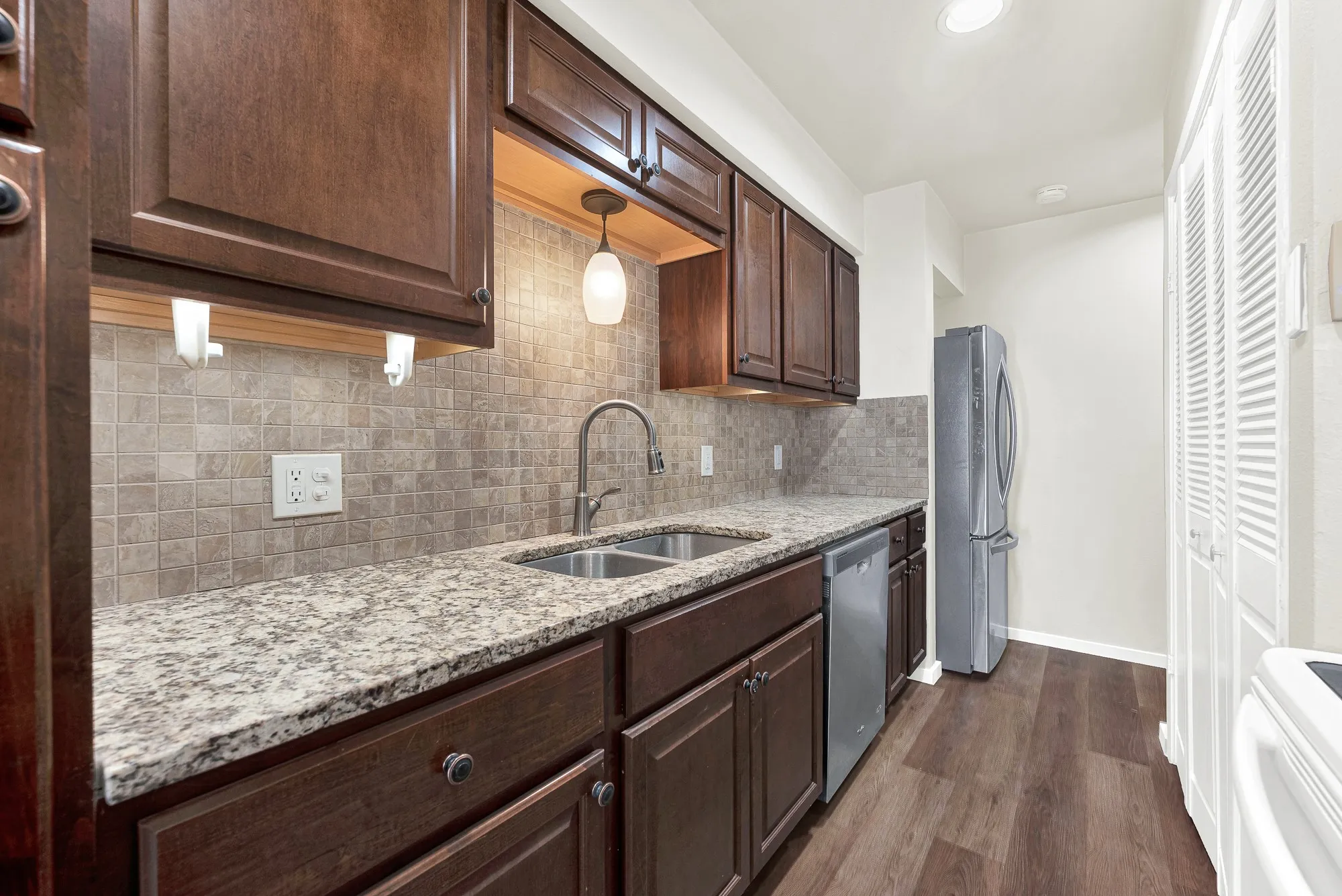 Wood-finish flooring, dark wood cabinetry, light-toned stone countertops, a tiled backsplash, and stainless steel appliances