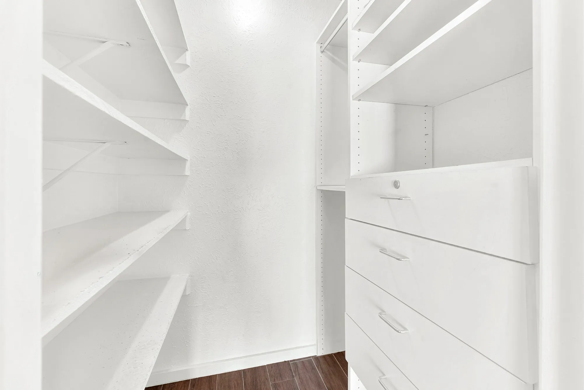 Custom closet system featuring multiple white shelves, integrated white drawers with silver hardware, and wood-finish flooring