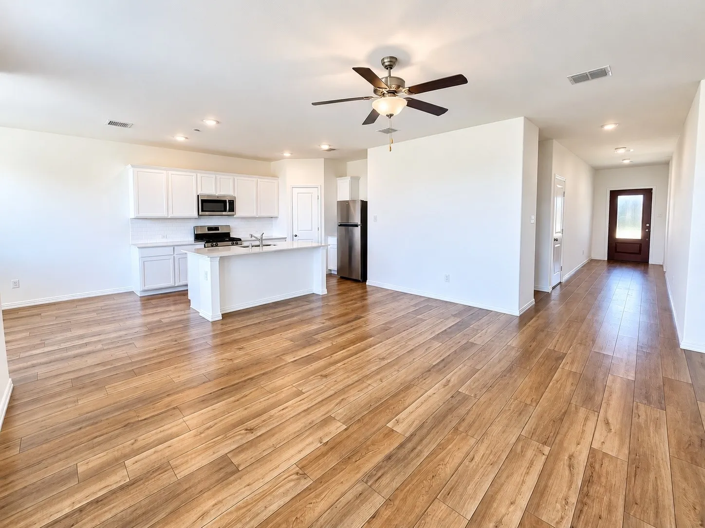 Open-concept layout featuring wood-finish flooring, a kitchen with white cabinetry and stainless steel appliances, a center island with a sink, and a ceiling fan with integrated lighting