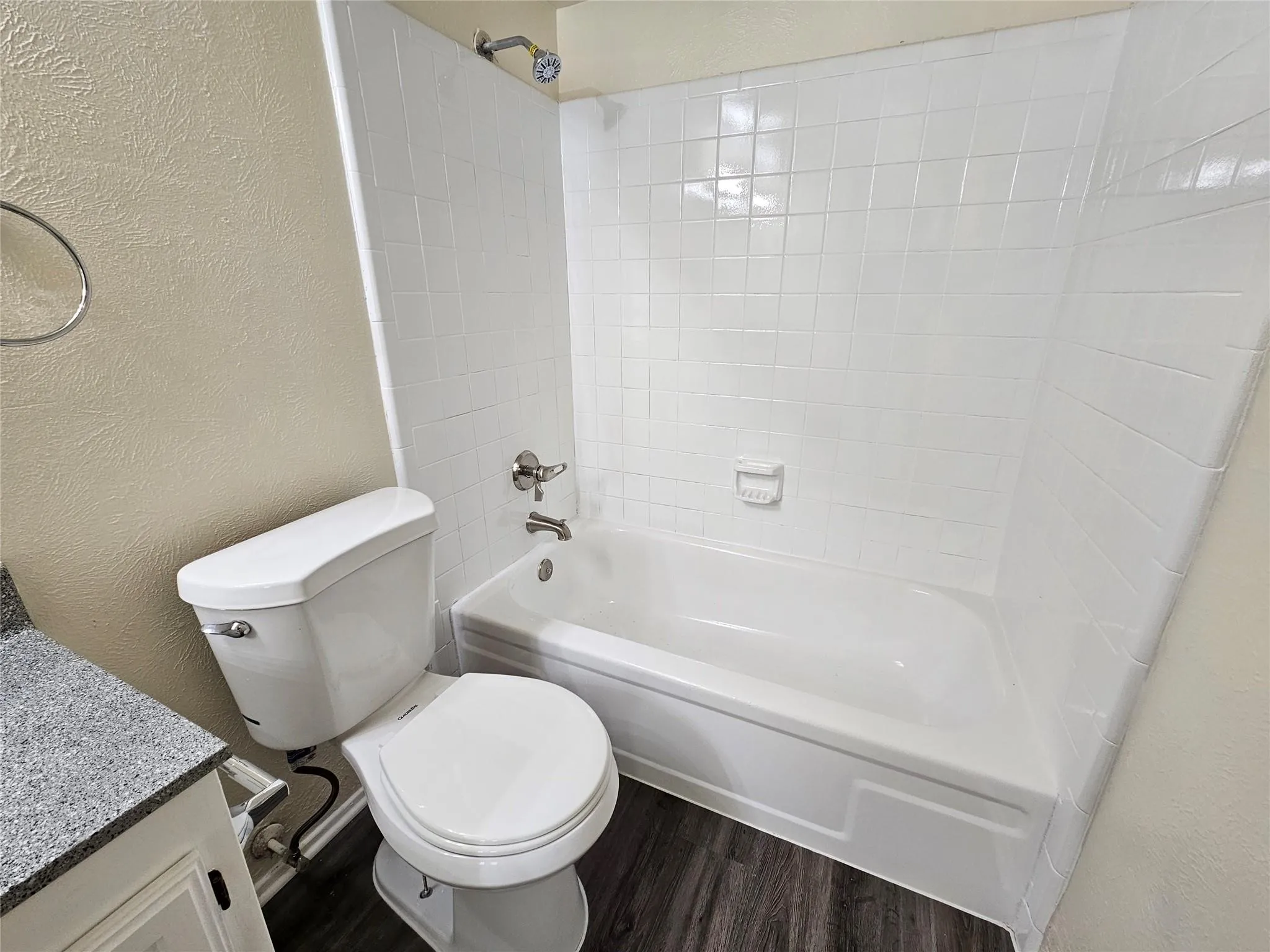 White porcelain bathtub and shower combination with tile surround, wood-finish flooring, and a white toilet