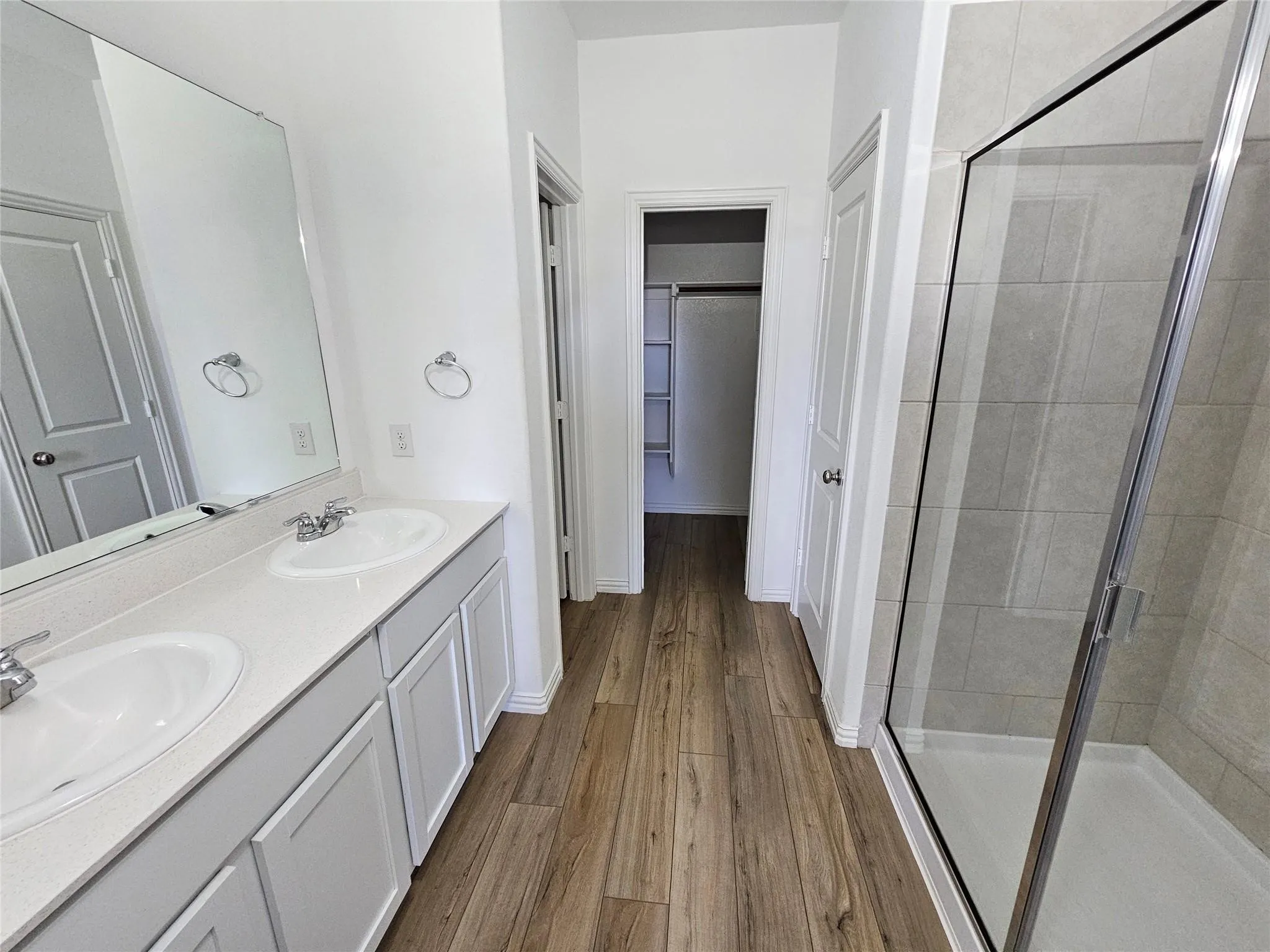 Dual vanity with integrated sinks, wood-finish flooring, glass-enclosed shower, and built-in closet shelving