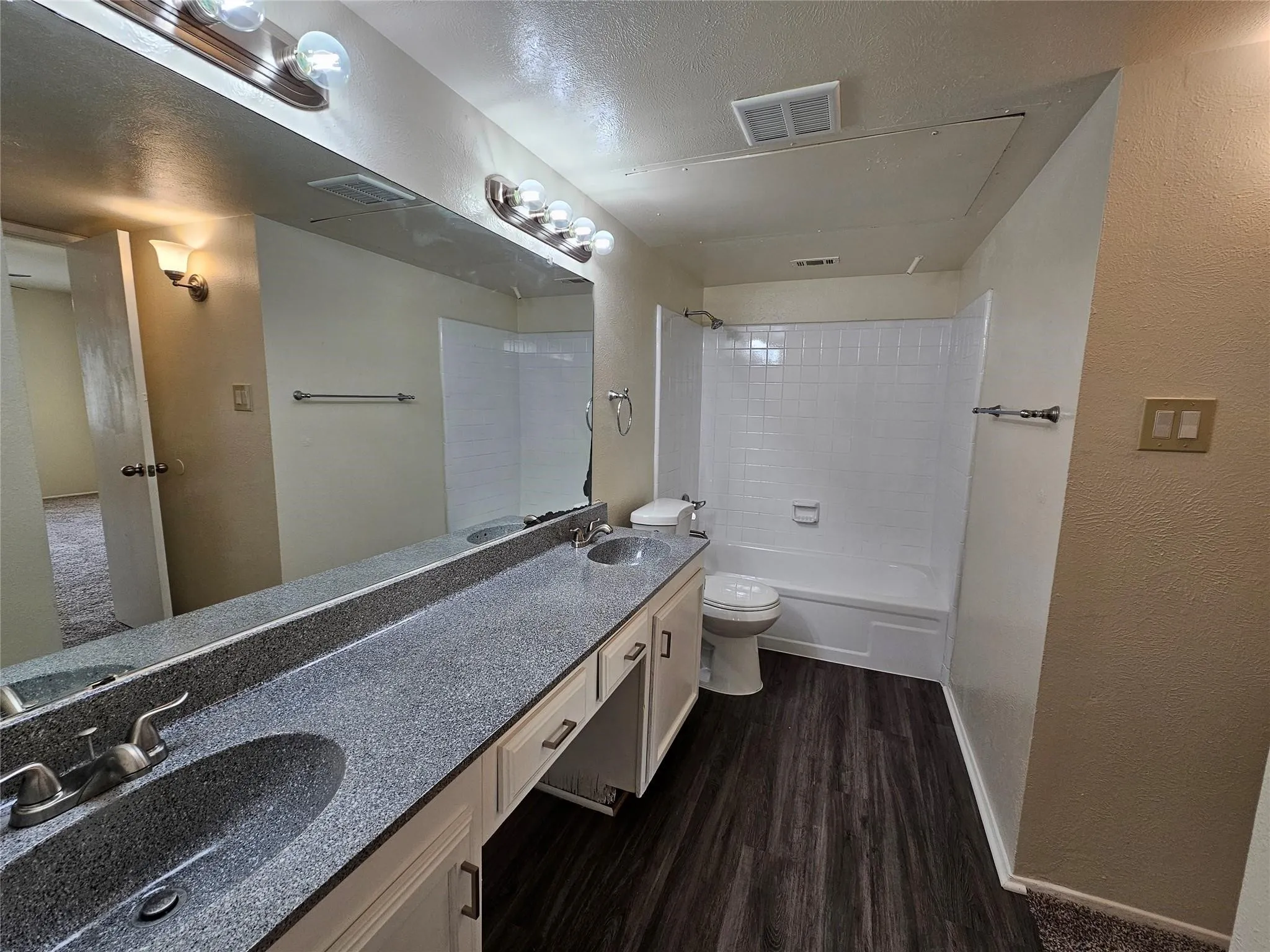 Dual vanity with integrated sinks, white cabinetry, dark wood-finish flooring, white tiled shower and tub combination, and wall-mounted light fixtures