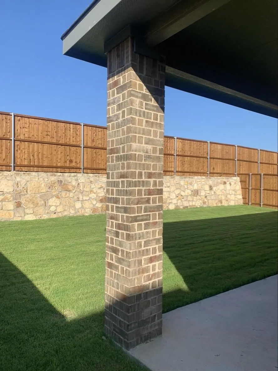 Expansive lawn with a masonry retaining wall, wood fencing, concrete patio, and a brick support column