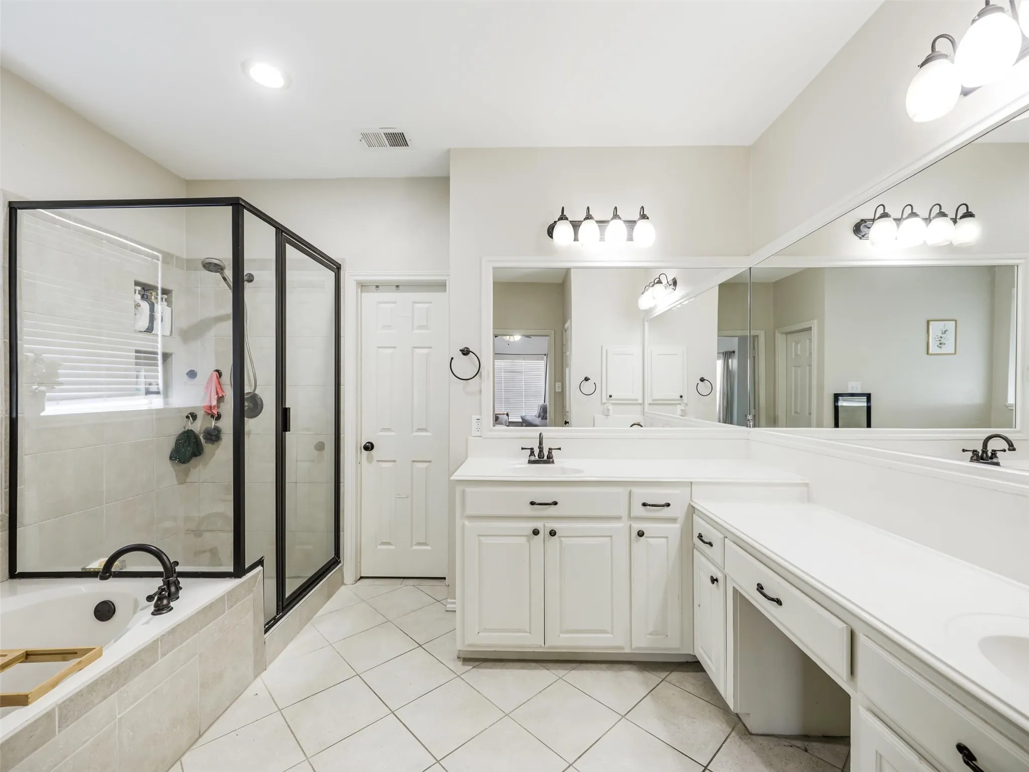 Black-framed glass shower enclosure, built-in soaking tub with black fixtures, dual white vanity with integrated sinks, expansive wall-mounted mirror, and tile flooring