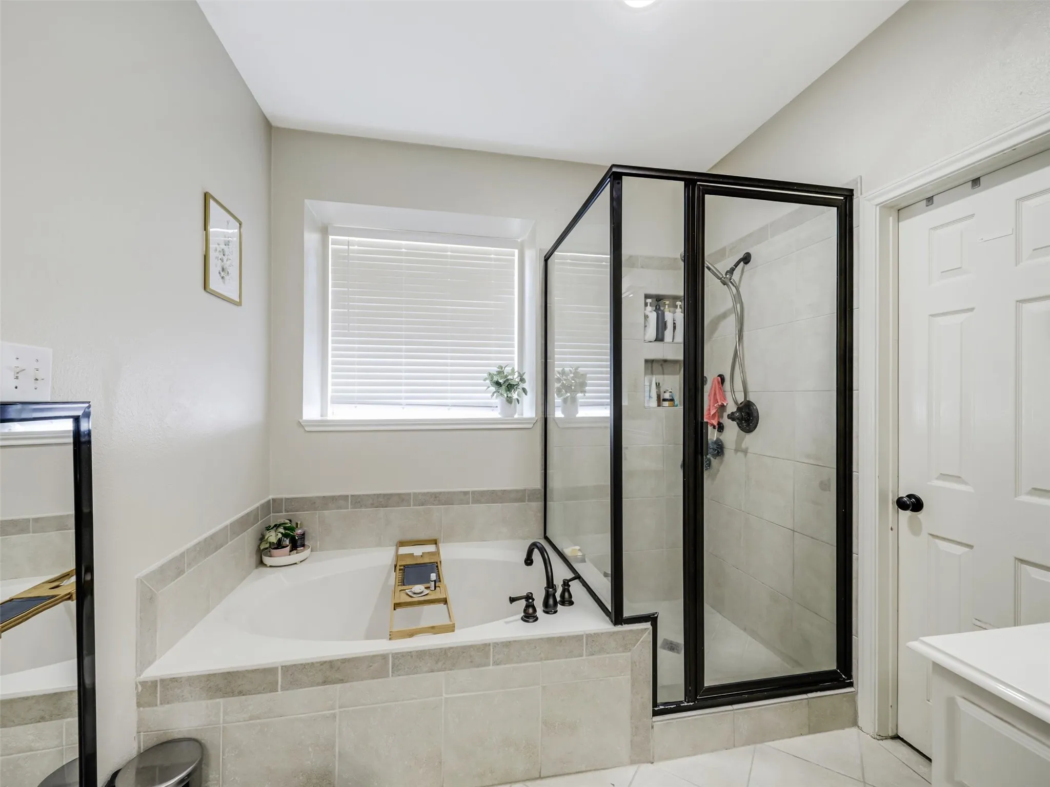 Built-in soaking tub with tiled surround, separate glass-enclosed shower with tiled walls, and window with blinds