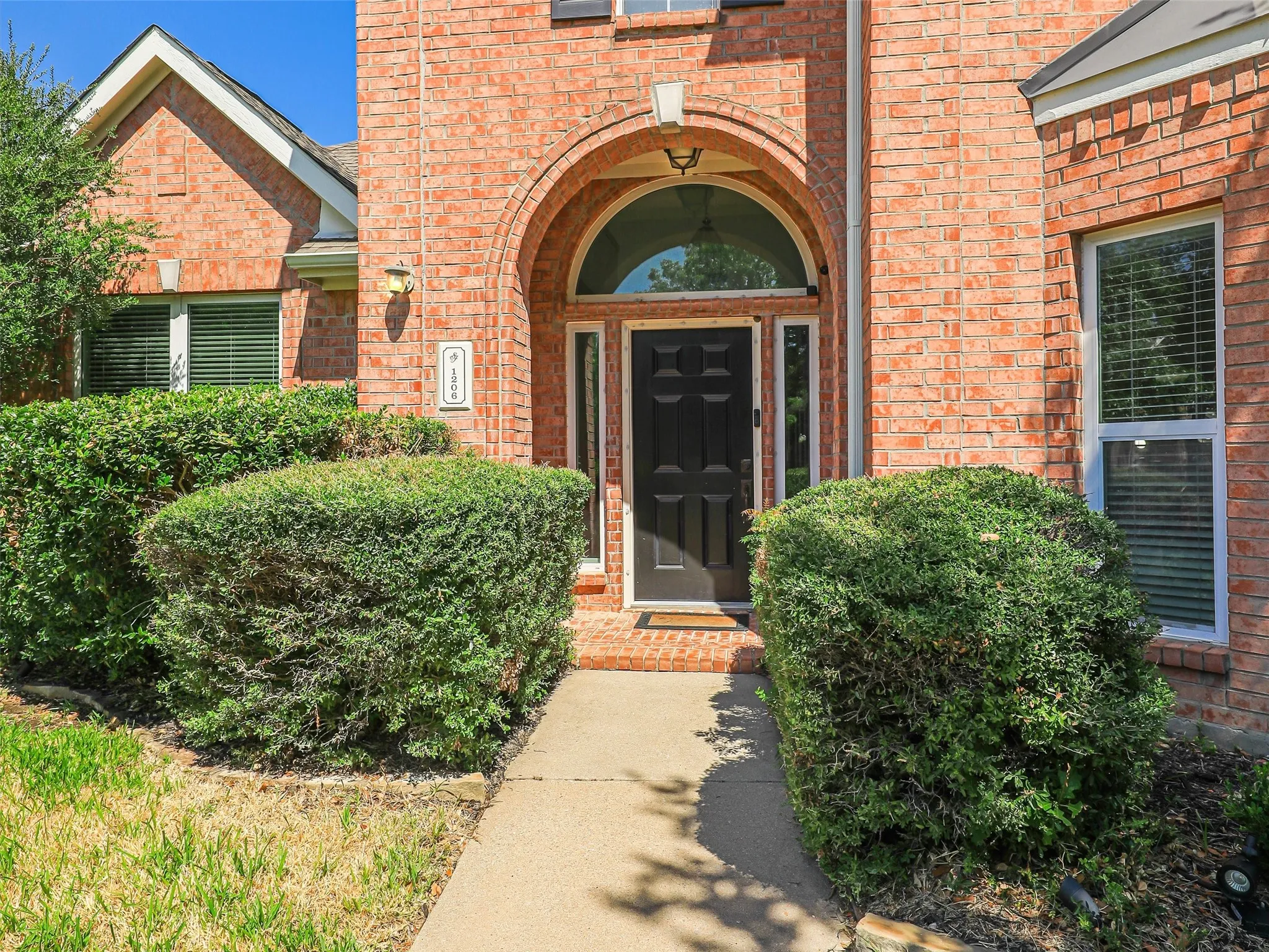 Brick exterior with arched entryway, black paneled door, and established shrubbery