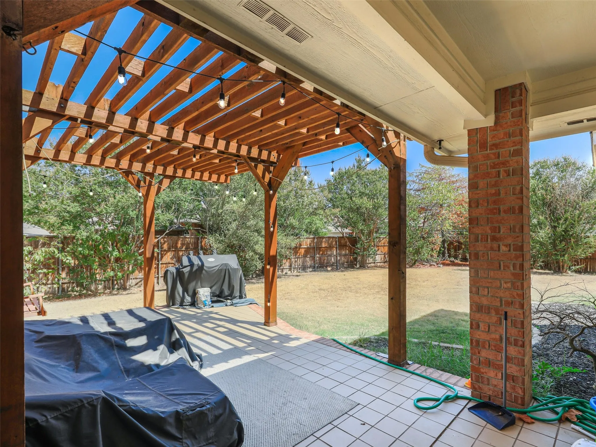 Expansive outdoor living area featuring a wood pergola with string lights, brick column accents, and a tiled patio surface