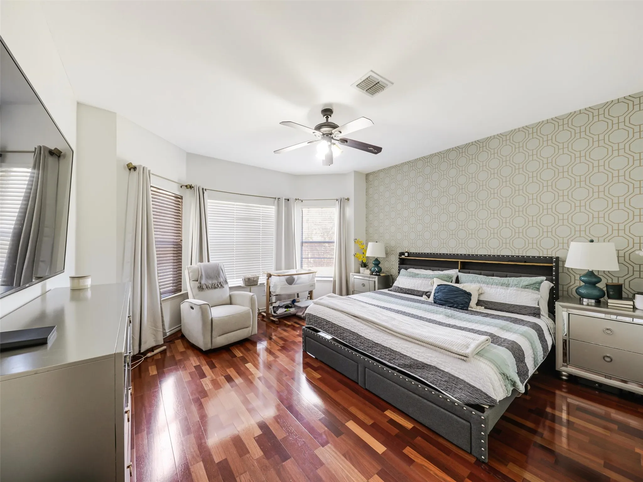 Hardwood flooring, bay window with blinds, ceiling fan, and accent wall