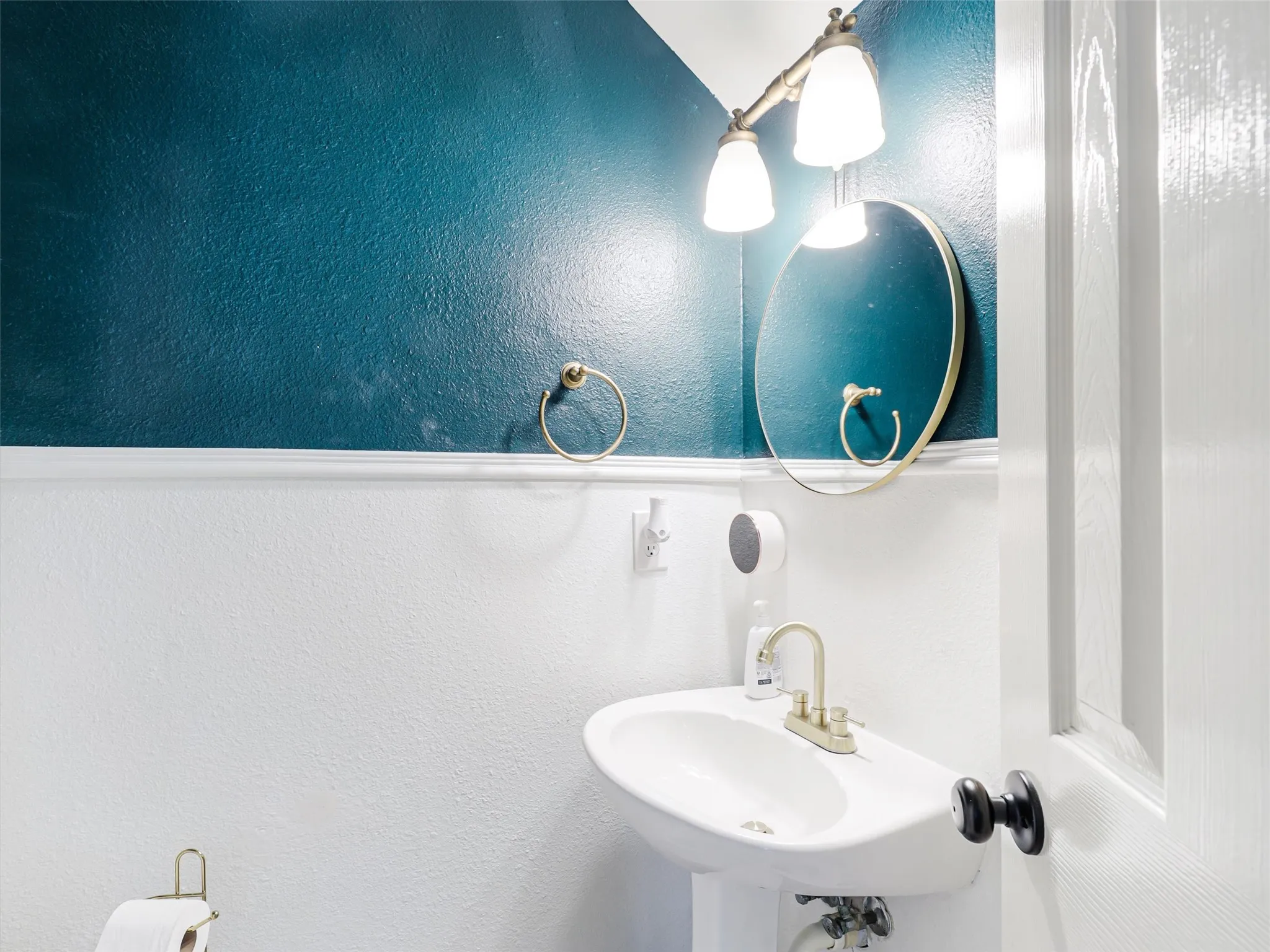 Pedestal sink with brass-finish faucet, circular mirror, and wall-mounted vanity light