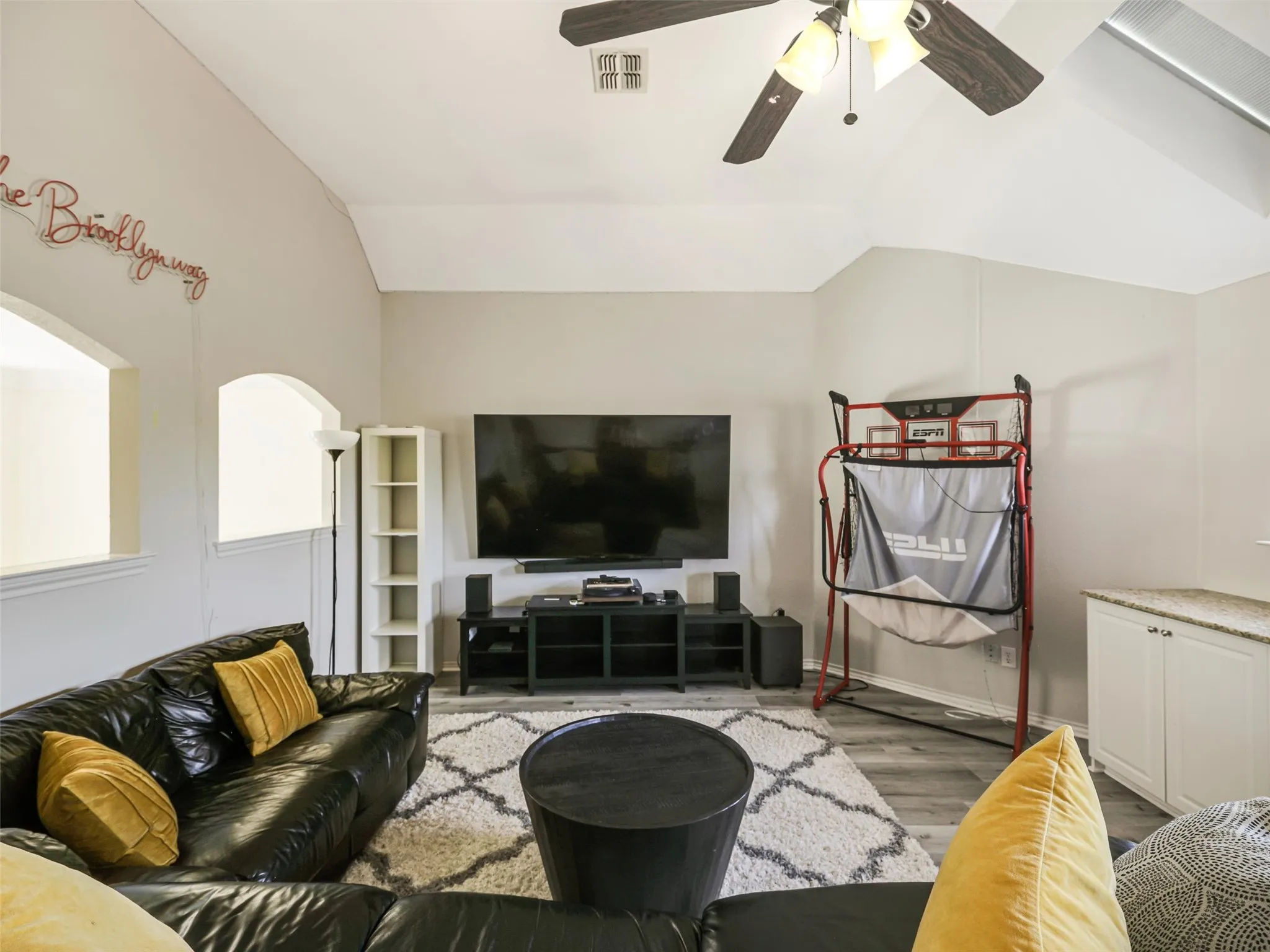 Vaulted ceiling with a ceiling fan light fixture, wood-finish flooring, built-in cabinetry with a stone-finish countertop, and arched openings