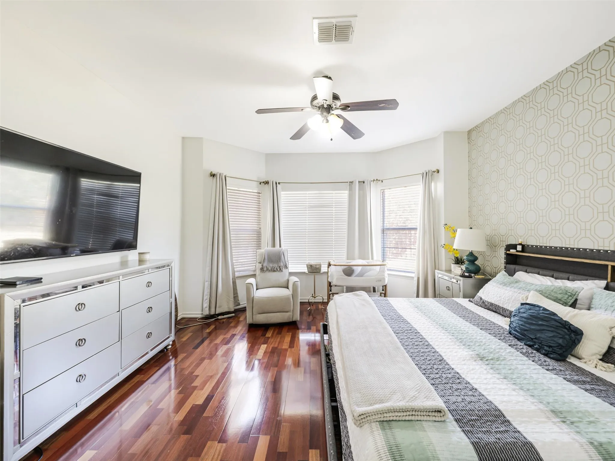 Rich wood-finish flooring, ceiling fan with integrated lighting, bay window, and accent wall with geometric pattern