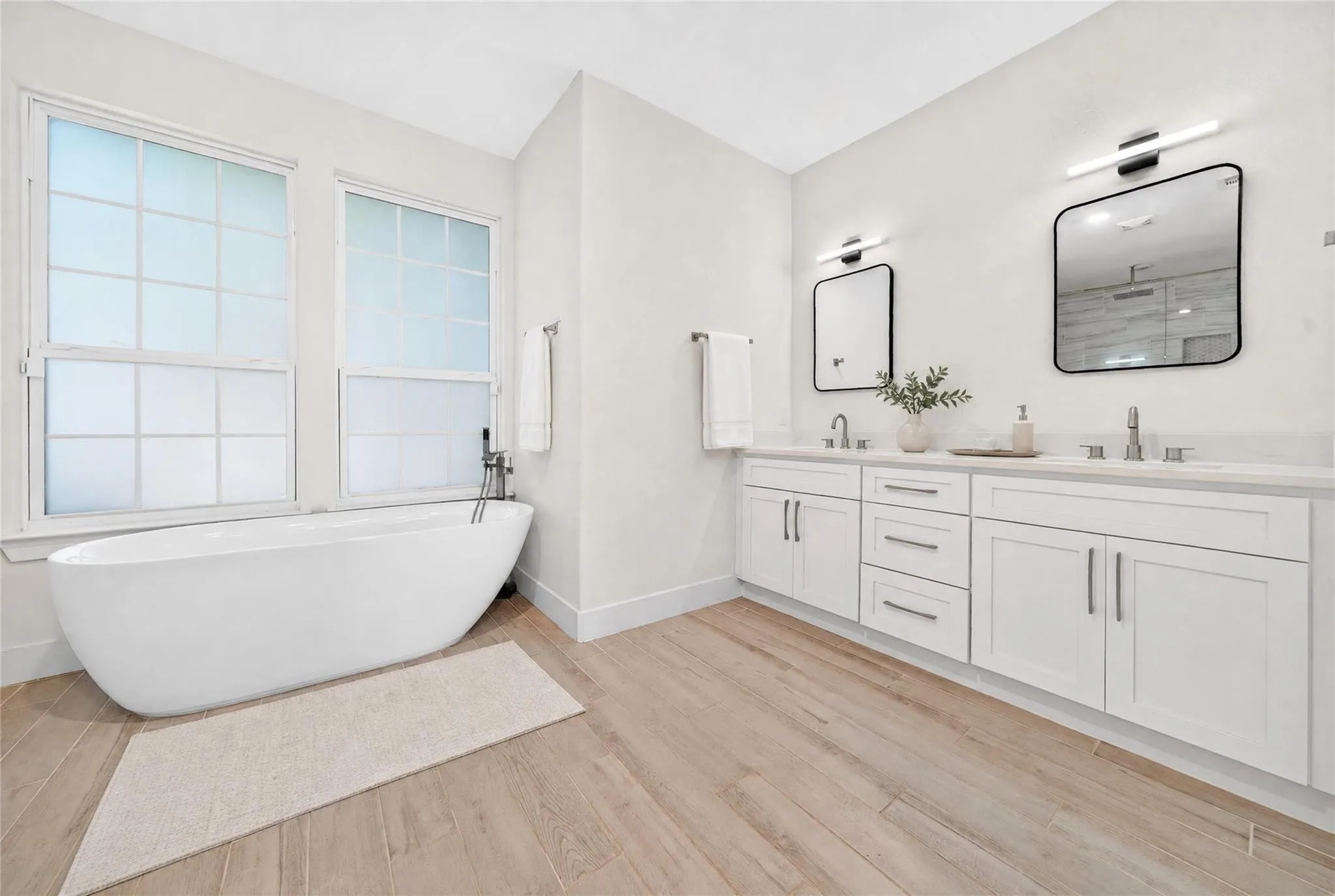 Freestanding soaking tub, dual windows with privacy glass, wood-finish flooring, dual vanity with white cabinetry, and wall-mounted light fixtures