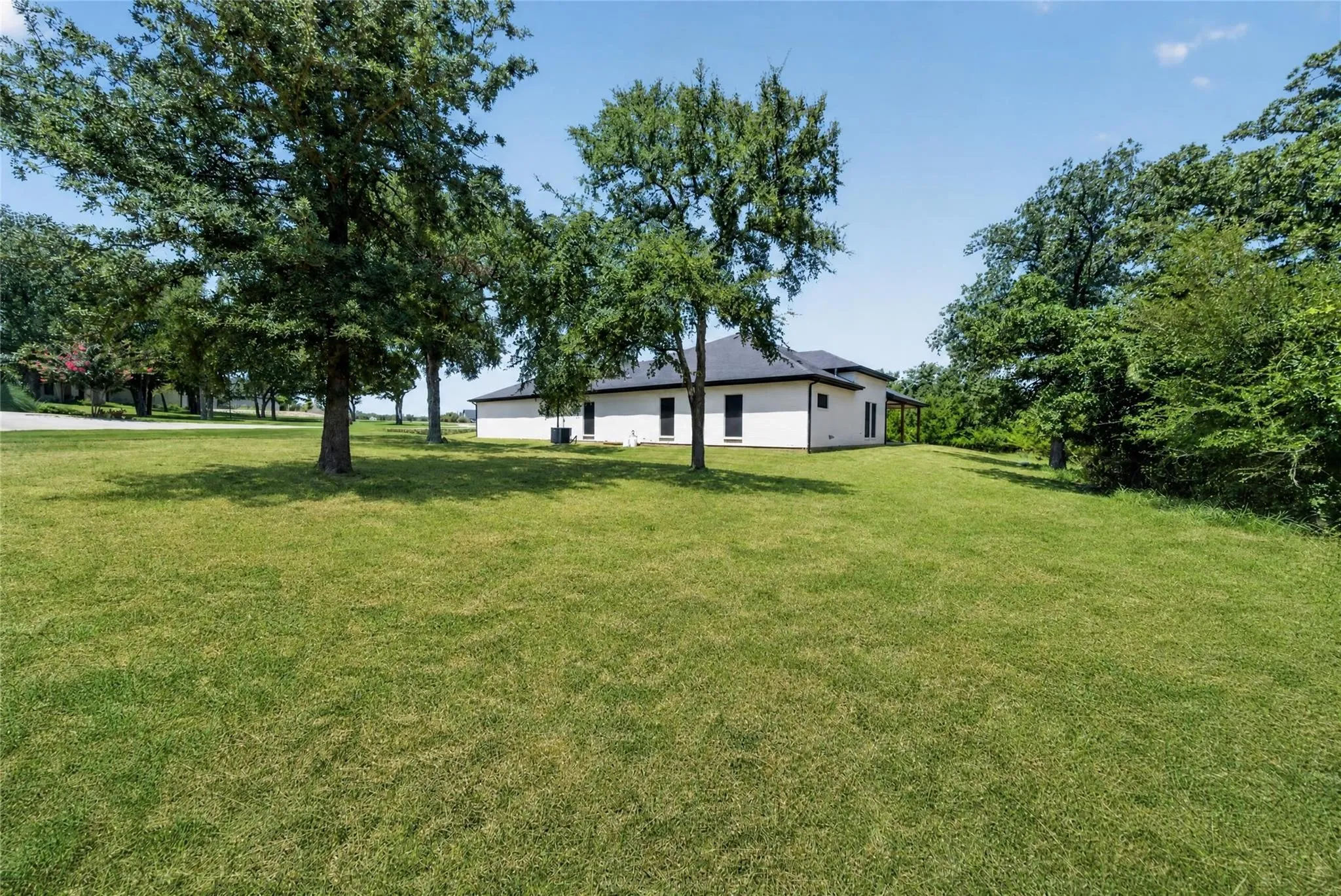 Expansive lawn area with mature trees, a single-story residence with a dark roof, and a covered porch