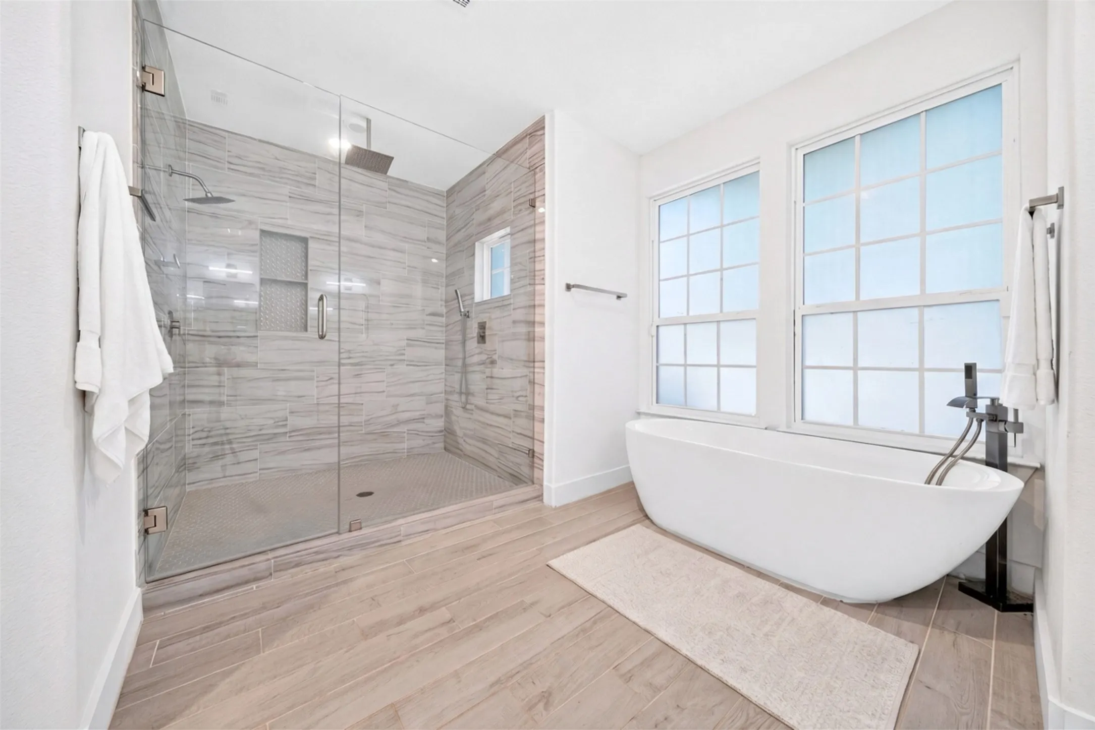 Spacious bathroom featuring a frameless glass shower enclosure, a freestanding soaking tub, wood-finish flooring, and white-framed windows