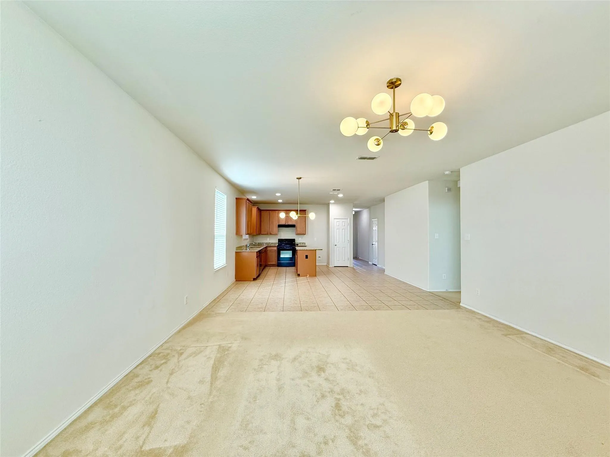 Open-concept living space with a modern chandelier, light-colored carpeting, and a kitchen featuring wood cabinetry and tile flooring