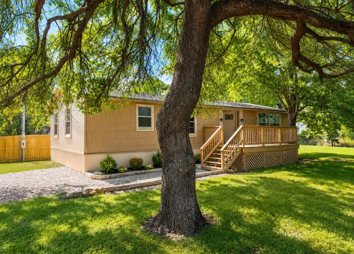 Exterior featuring tan siding, a wooden deck with railing, a gravel driveway, and a fenced perimeter