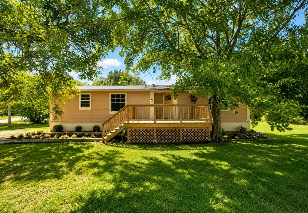 Manicured lawn with mature trees and a front porch featuring a wooden railing and lattice skirting
