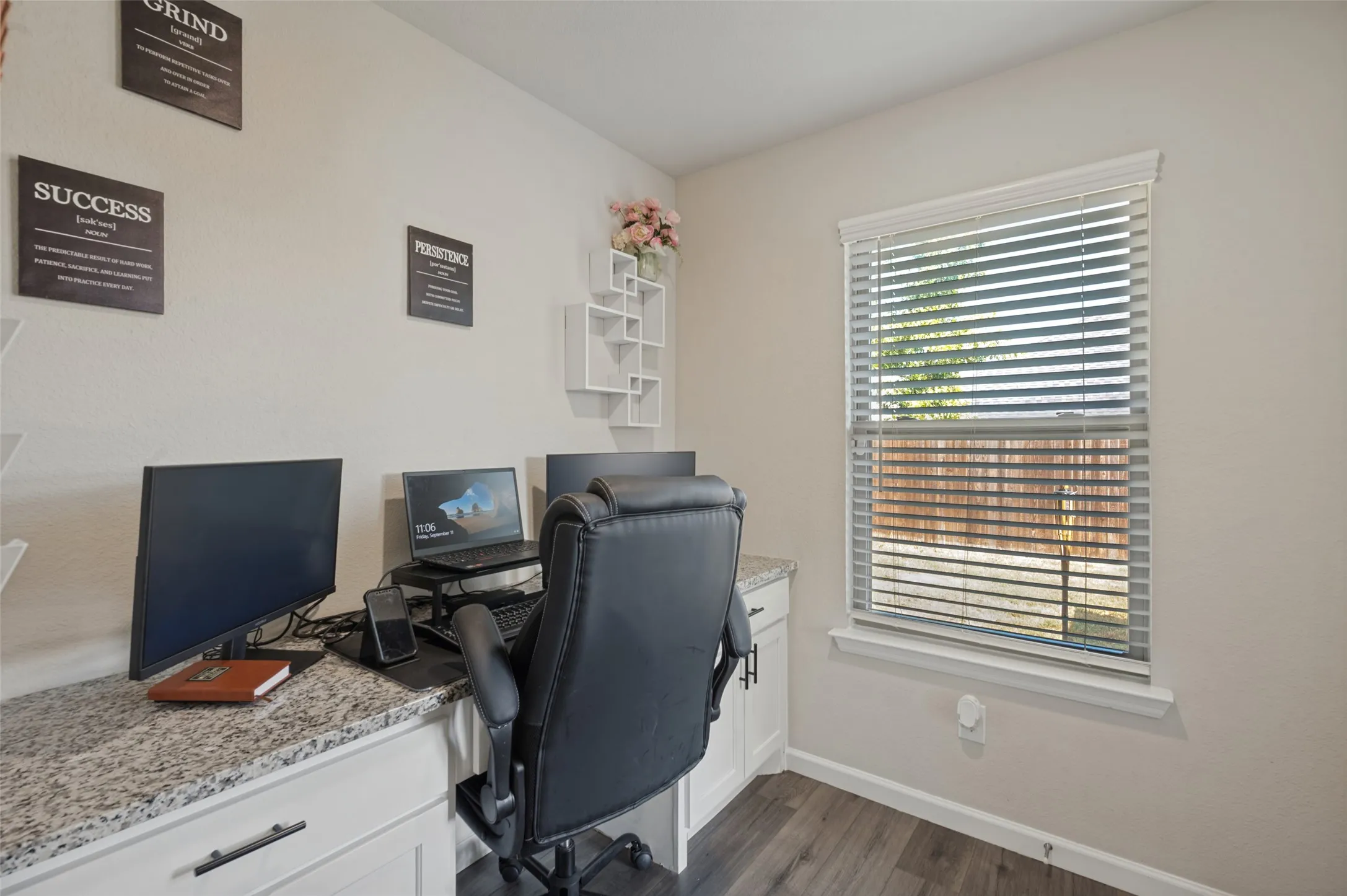 Dedicated home office featuring a wood-finish desk with quartz countertop, ergonomic seating, dual monitor setup capability, and abundant natural light from a large window with plantation shutters
