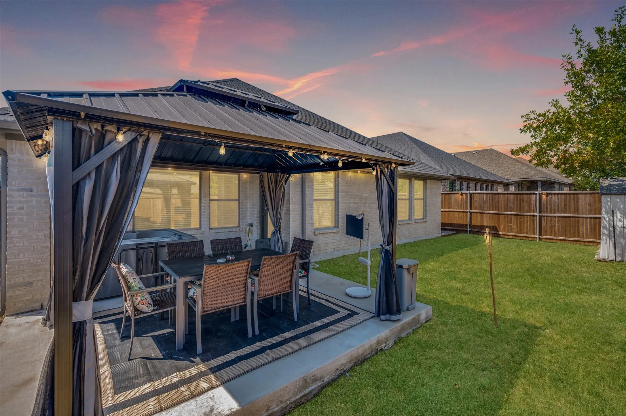 Covered patio with string lighting, surrounded by a fenced backyard featuring a grassy area and brick exterior