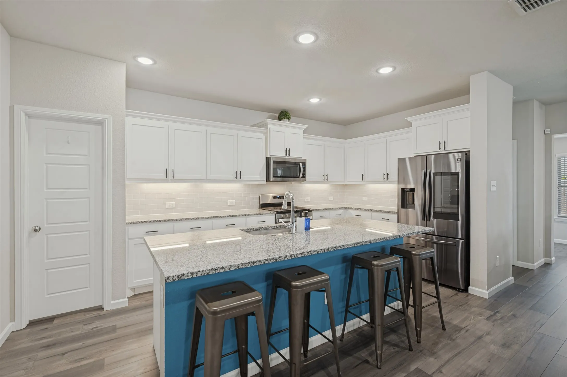 Kitchen featuring an expansive island with a blue base and light-colored countertop, white upper and lower cabinetry, stainless steel appliances, and wood-finish flooring