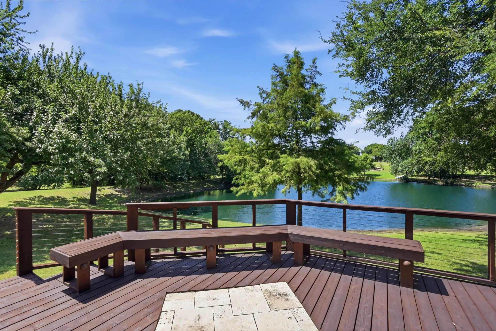 Wood-finish deck with built-in bench seating, cable railing, and stone-finish flooring accent, overlooking a body of water and expansive green space