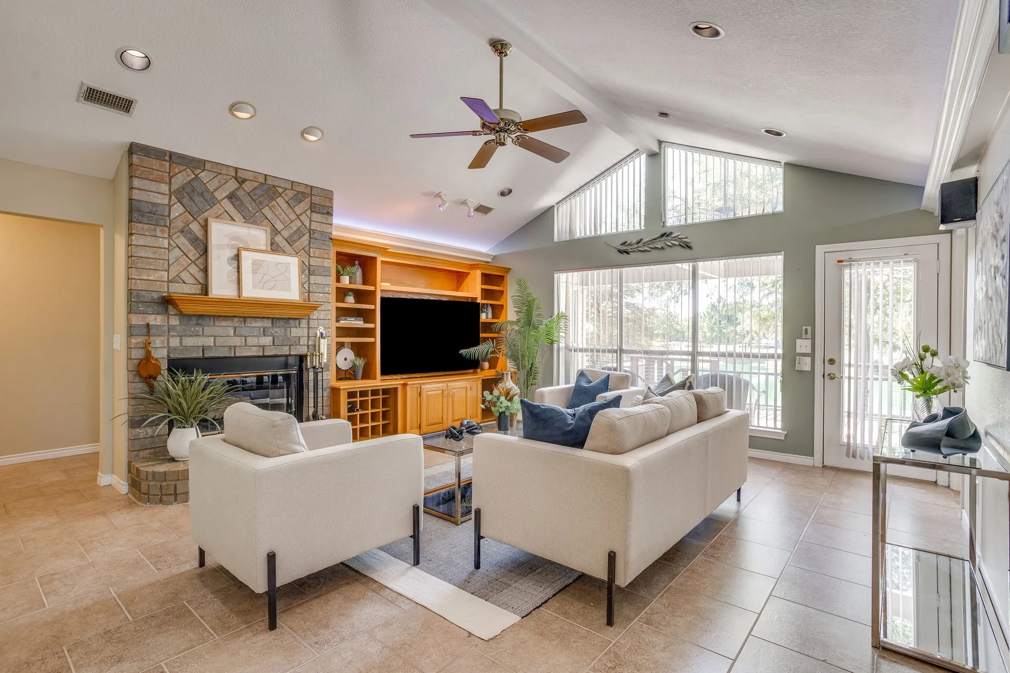Vaulted ceiling with recessed lighting, brick fireplace with wood mantel, built-in entertainment center with shelving, large windows, and tile flooring