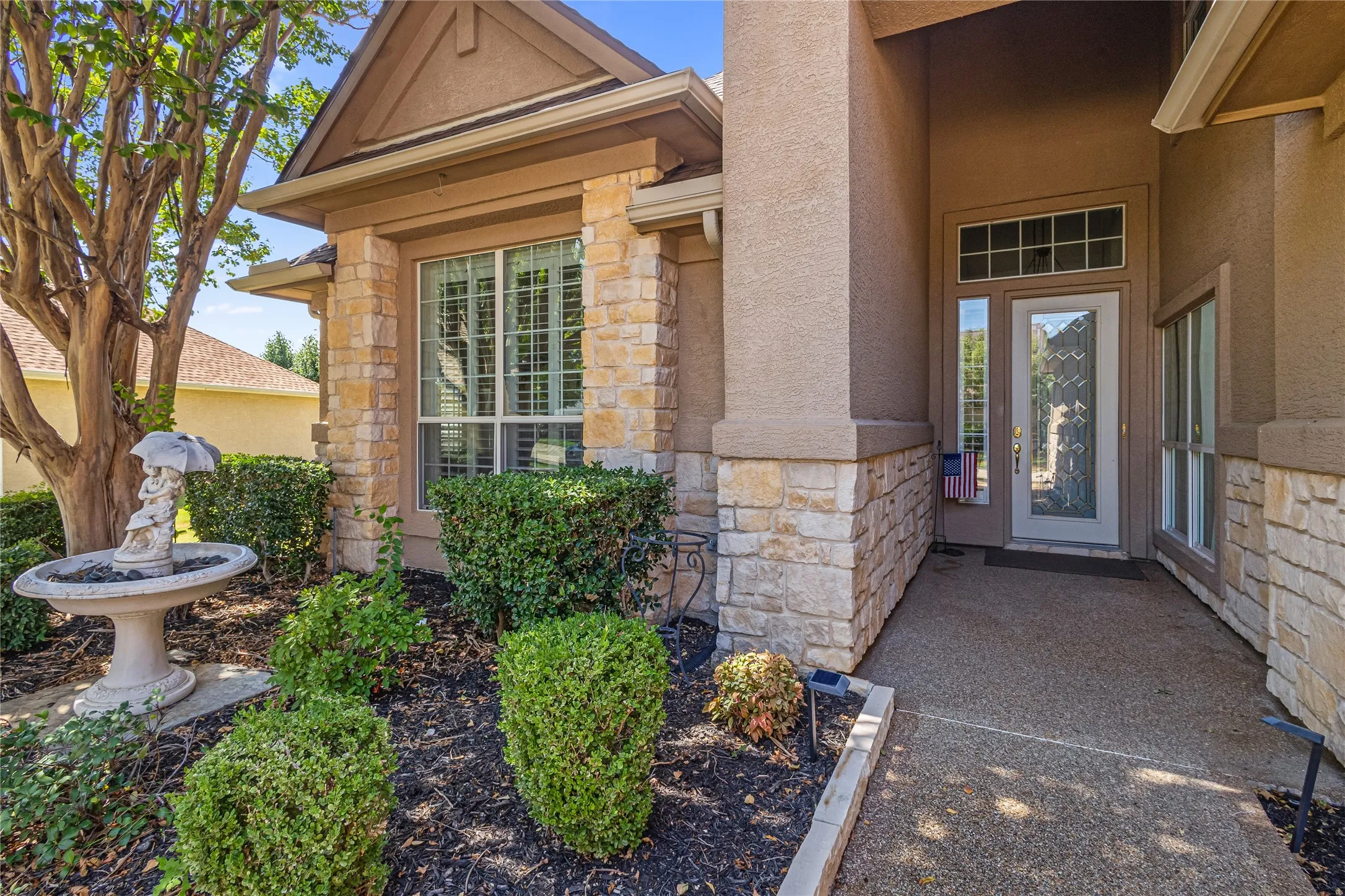 Stucco and stone exterior, mature tree, established landscaping, decorative fountain, and an entryway with a transom window