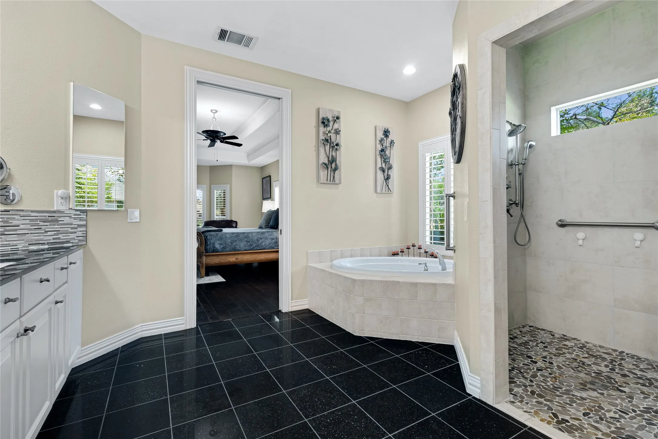 Black tile flooring, built-in soaking tub, walk-in shower with pebble floor, white vanity with stone countertop, and tile backsplash