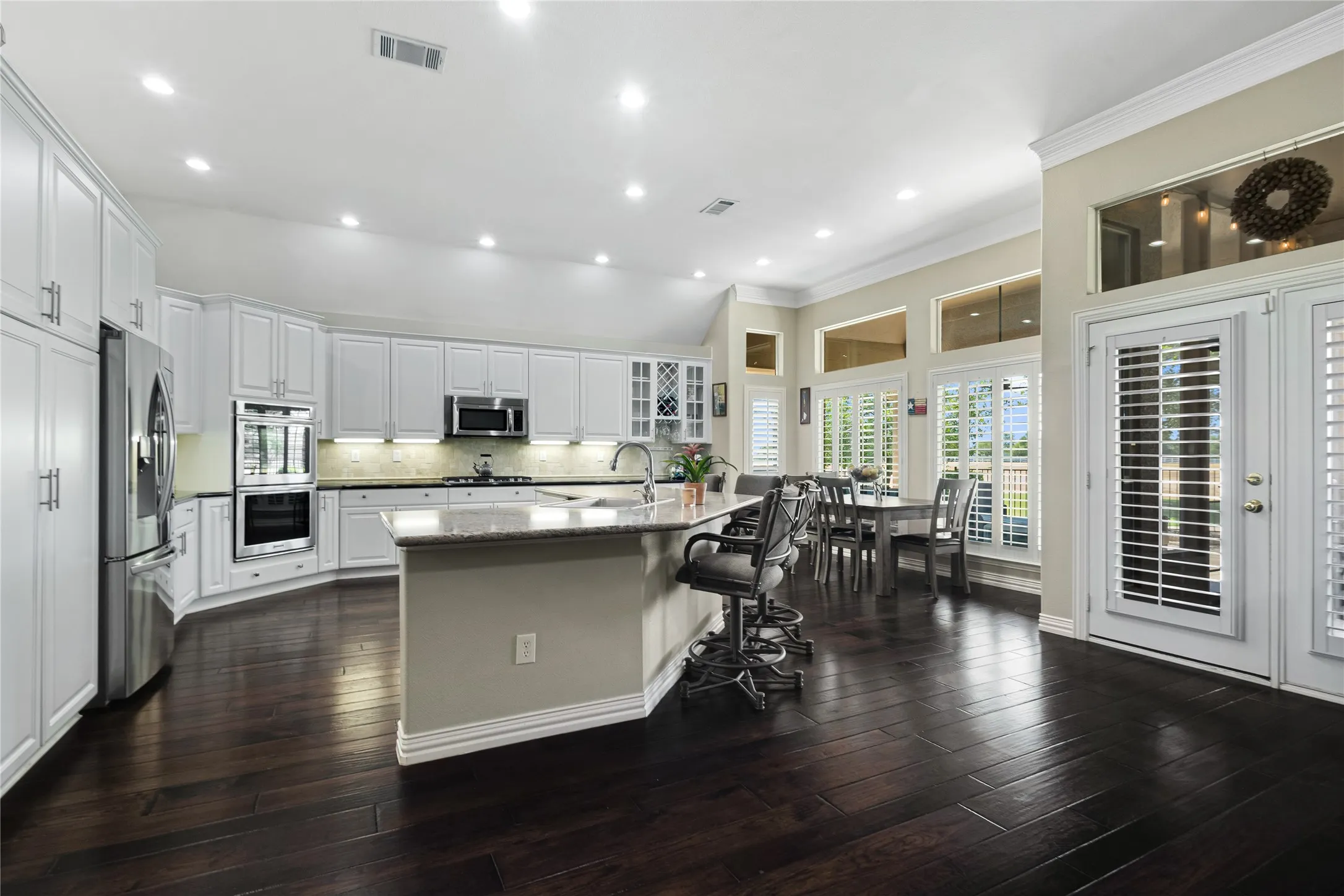 Gourmet kitchen with white cabinetry, stainless steel appliances, a large island with counter seating, dark wood-finish flooring, and multiple windows with plantation shutters