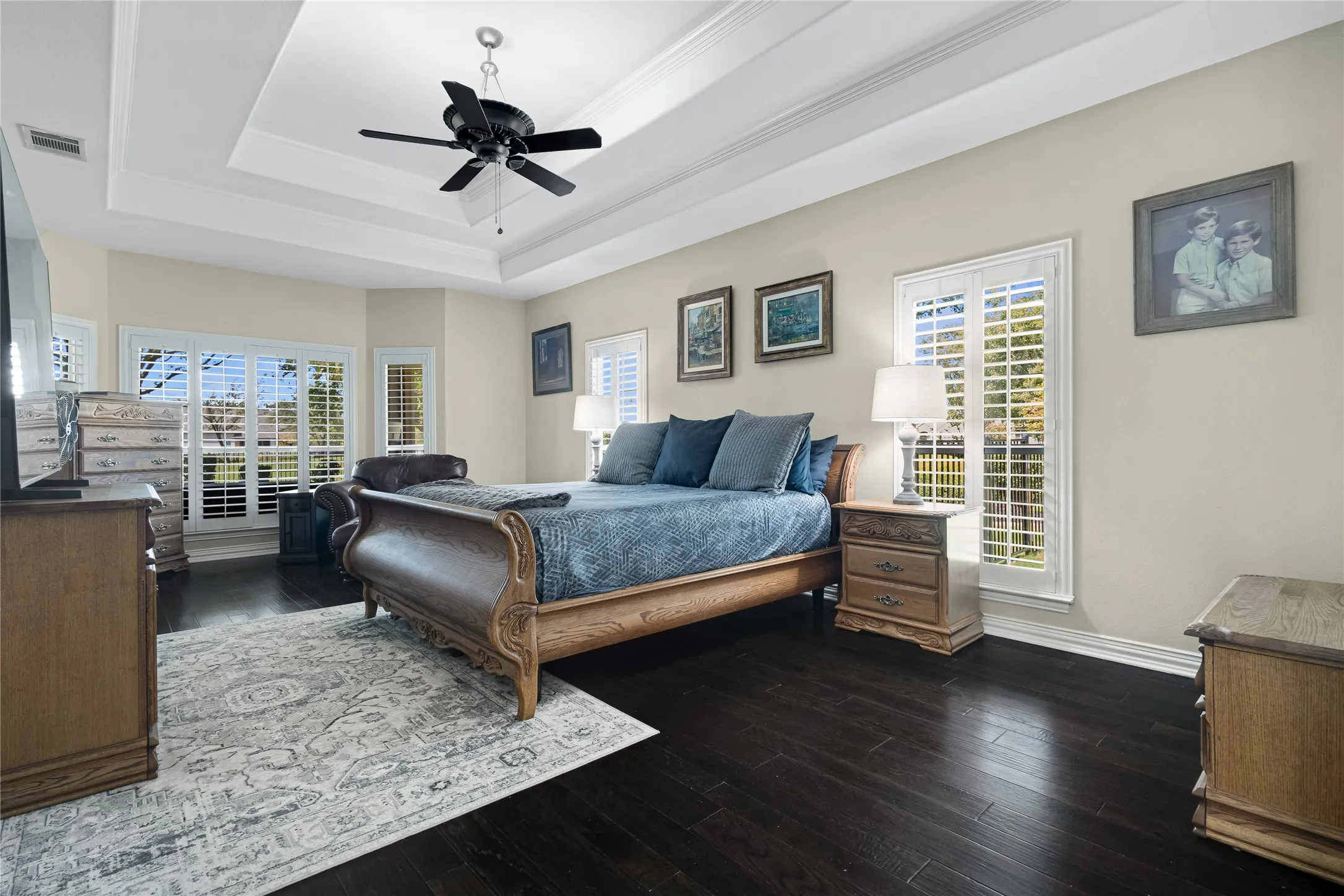 Tray ceiling with crown molding, dark wood-finish flooring, multiple windows with white plantation shutters, and a ceiling fan with light fixture