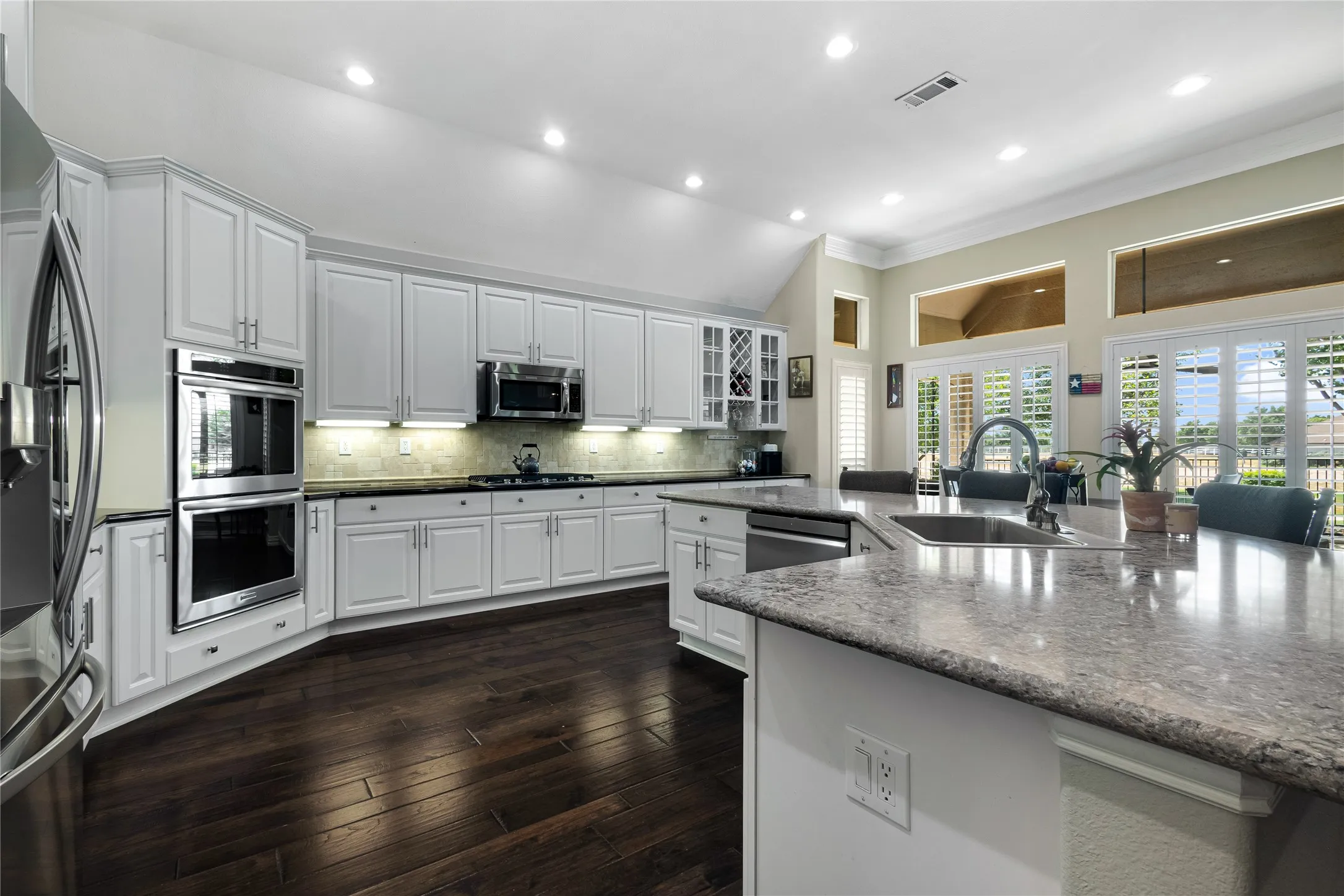 Spacious kitchen featuring white cabinetry, dark wood-finish flooring, stainless steel appliances including double ovens, and a large central island with an integrated sink