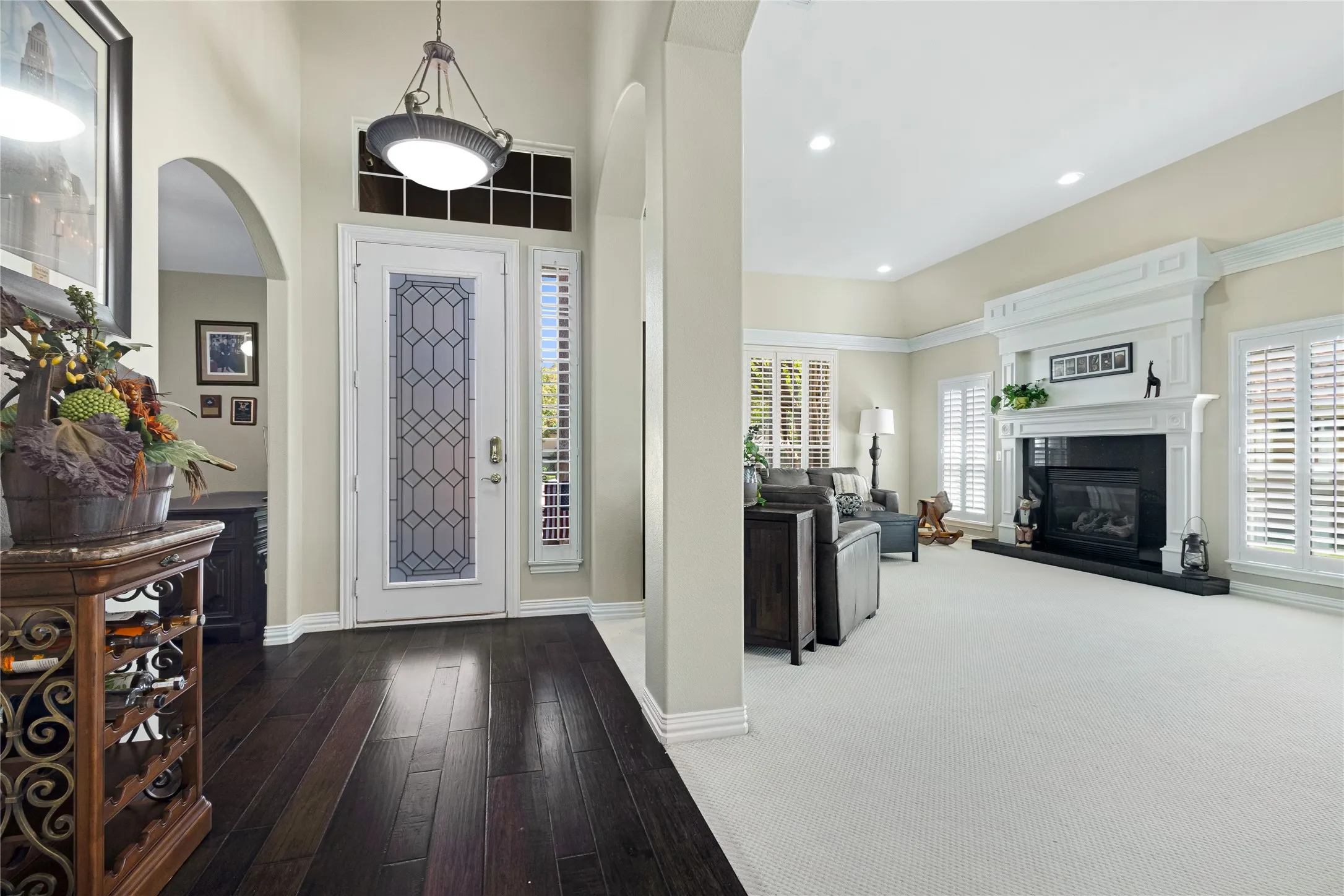 Hardwood flooring extending from the entryway, fireplace with substantial mantel, recessed lighting, arched doorway, and plantation shutters