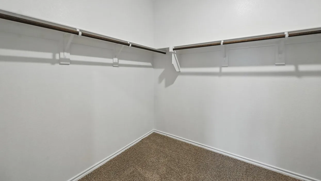 Walk-in closet featuring light brown carpet flooring, white painted walls, and an L-shaped clothes rod with support brackets