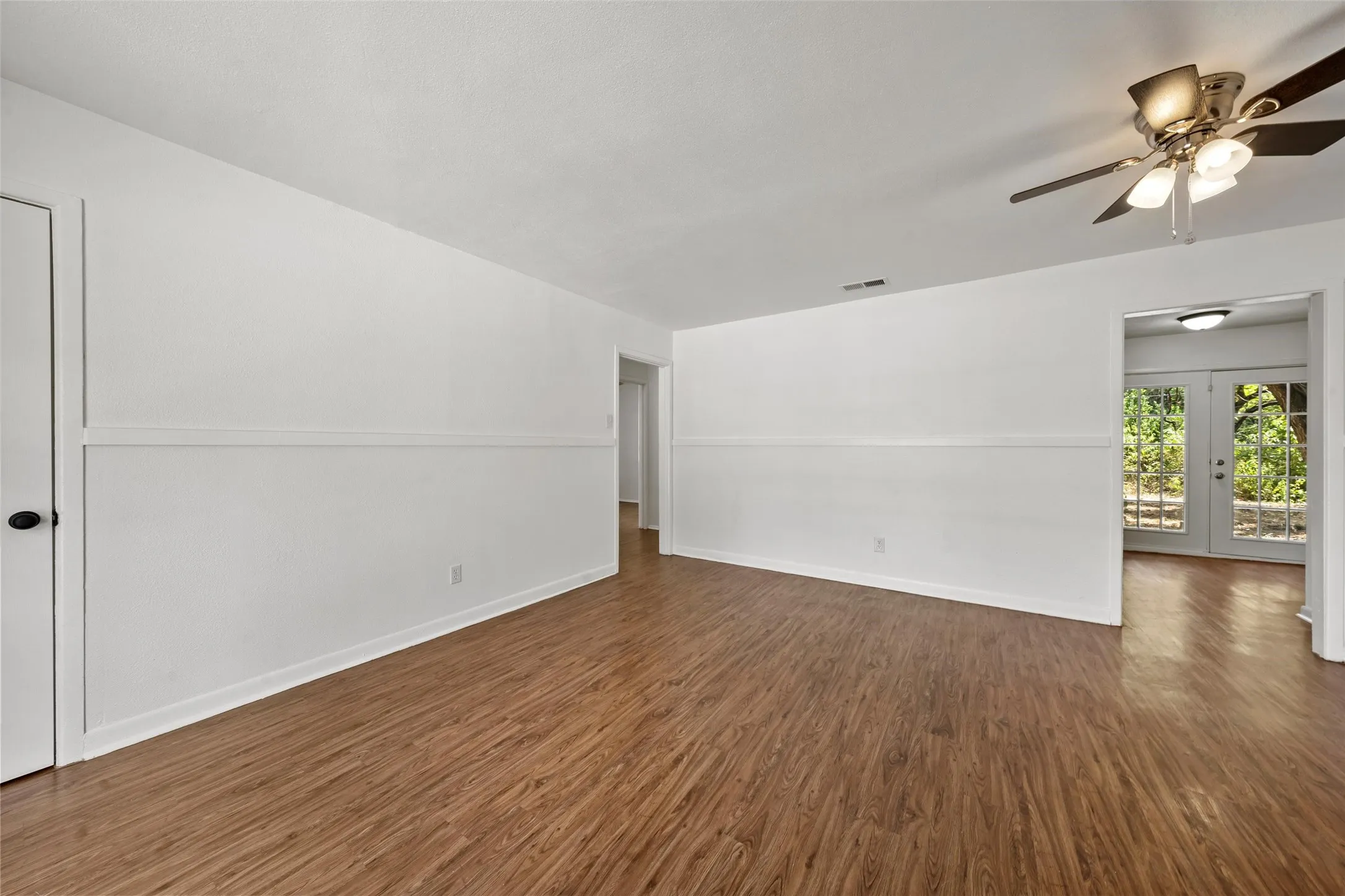 Wood-finish flooring, white walls with chair rail molding, ceiling fan with integrated lighting, and a doorway leading to outdoor access with double glass-paneled doors
