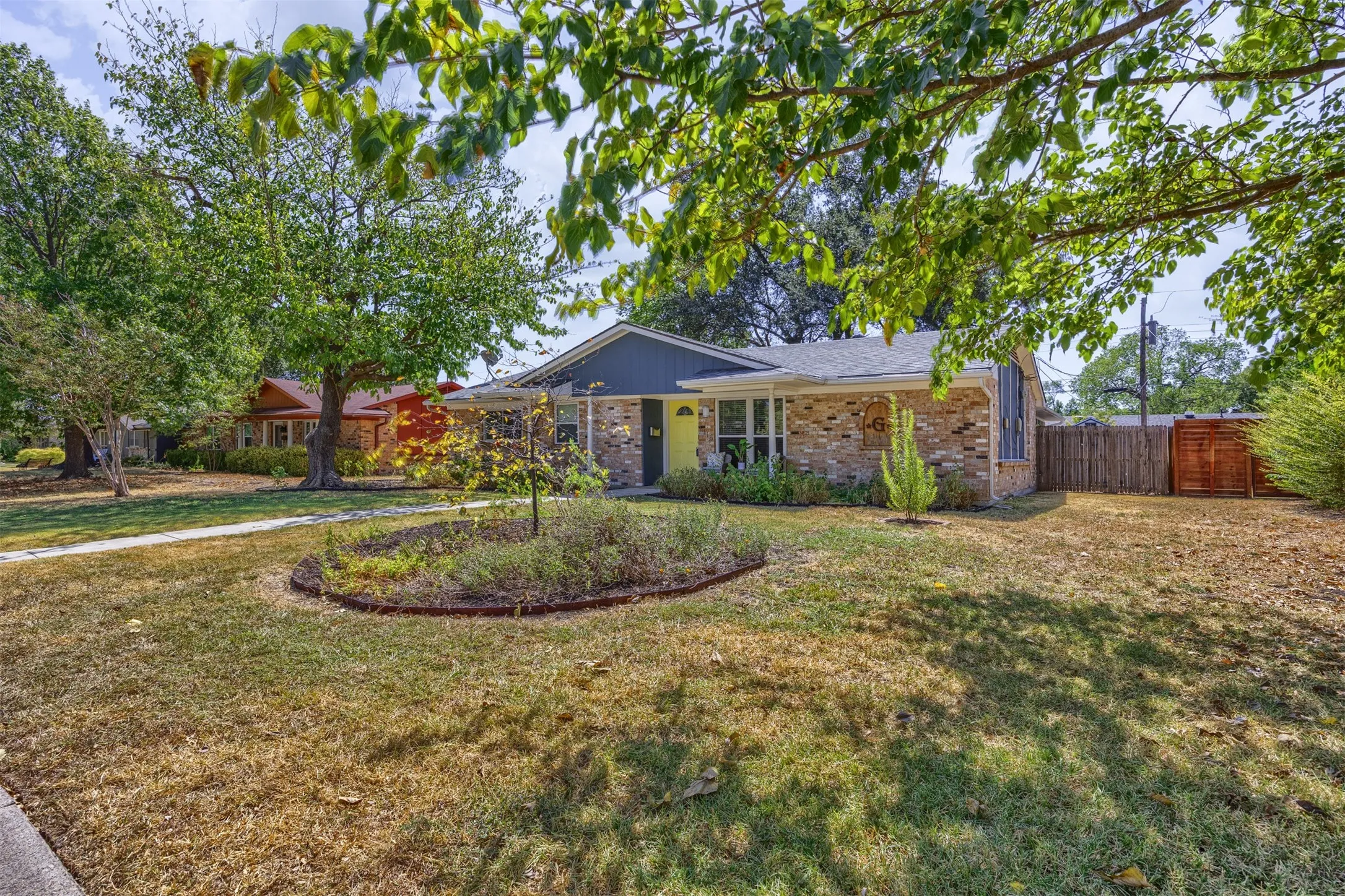 Brick exterior with a contrasting façade, a central landscaped garden bed, mature trees, and a privacy fence