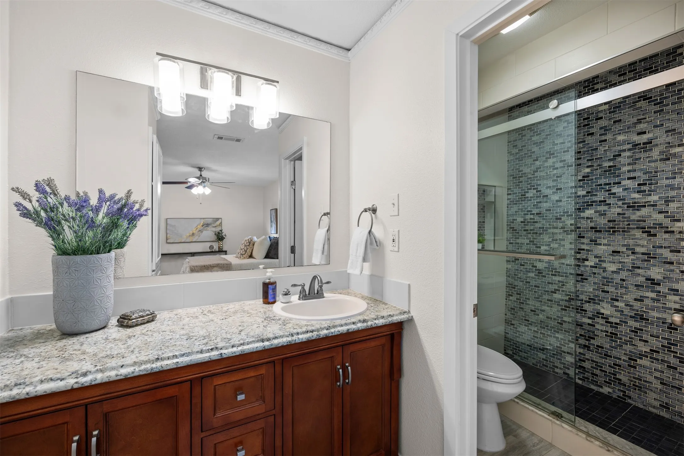 Granite countertop vanity with an undermount sink, wood-finish cabinetry, a large mirror, and an adjacent shower with glass enclosure and mosaic tile