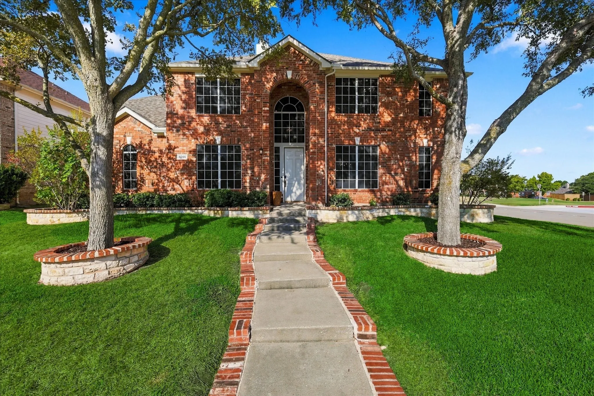 Two-story brick exterior, arched entryway, symmetrical window placement, concrete pathway with brick edging, and mature trees with stone and brick planters