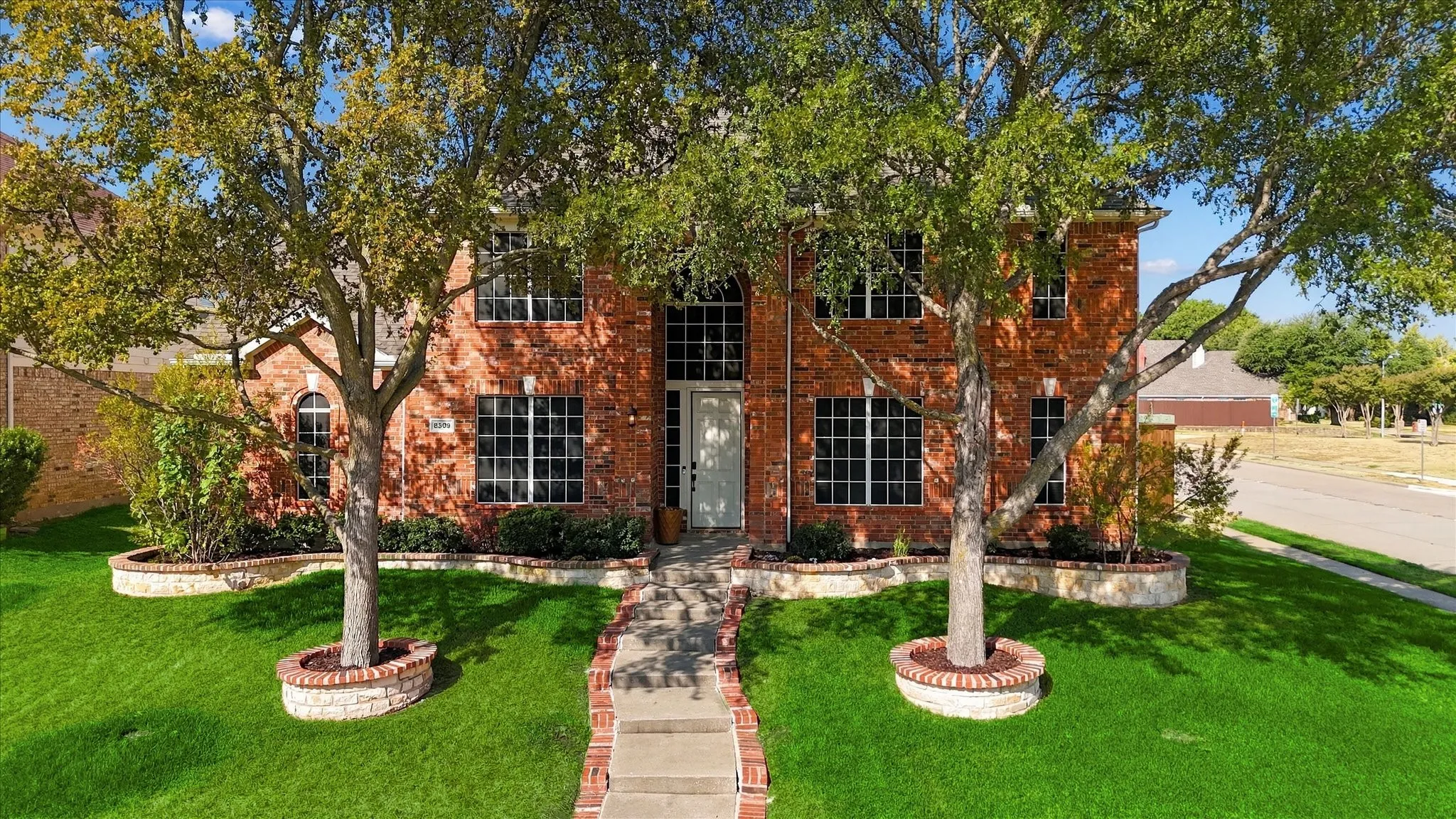 Charming brick residence featuring a symmetrical facade with multiple tall windows, mature shade trees, and a manicured lawn with defined planting beds