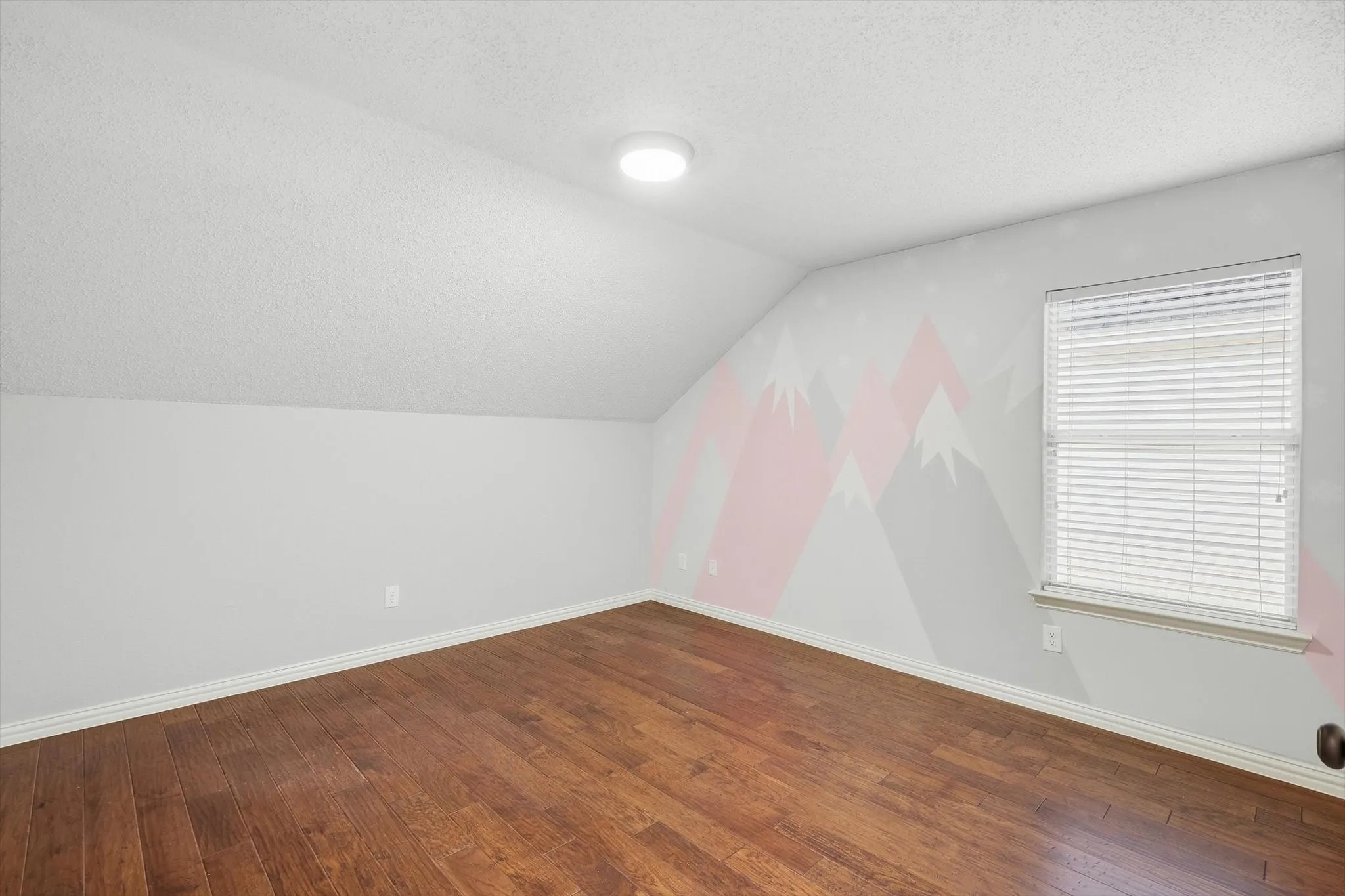 Wood-finish flooring with a sloped ceiling and a window featuring blinds