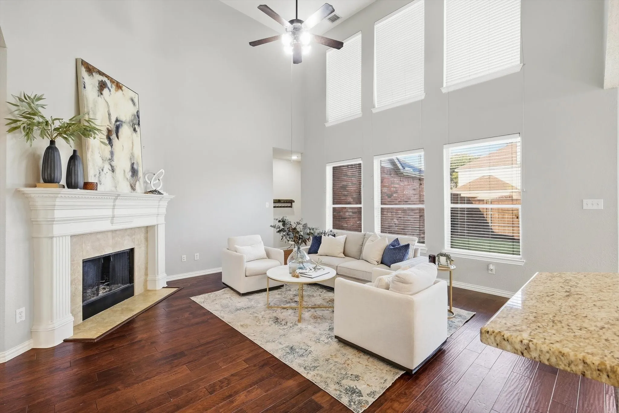 Hardwood flooring, high vaulted ceilings, large windows with blinds, a ceiling fan, and a stone-front fireplace with a mantel
