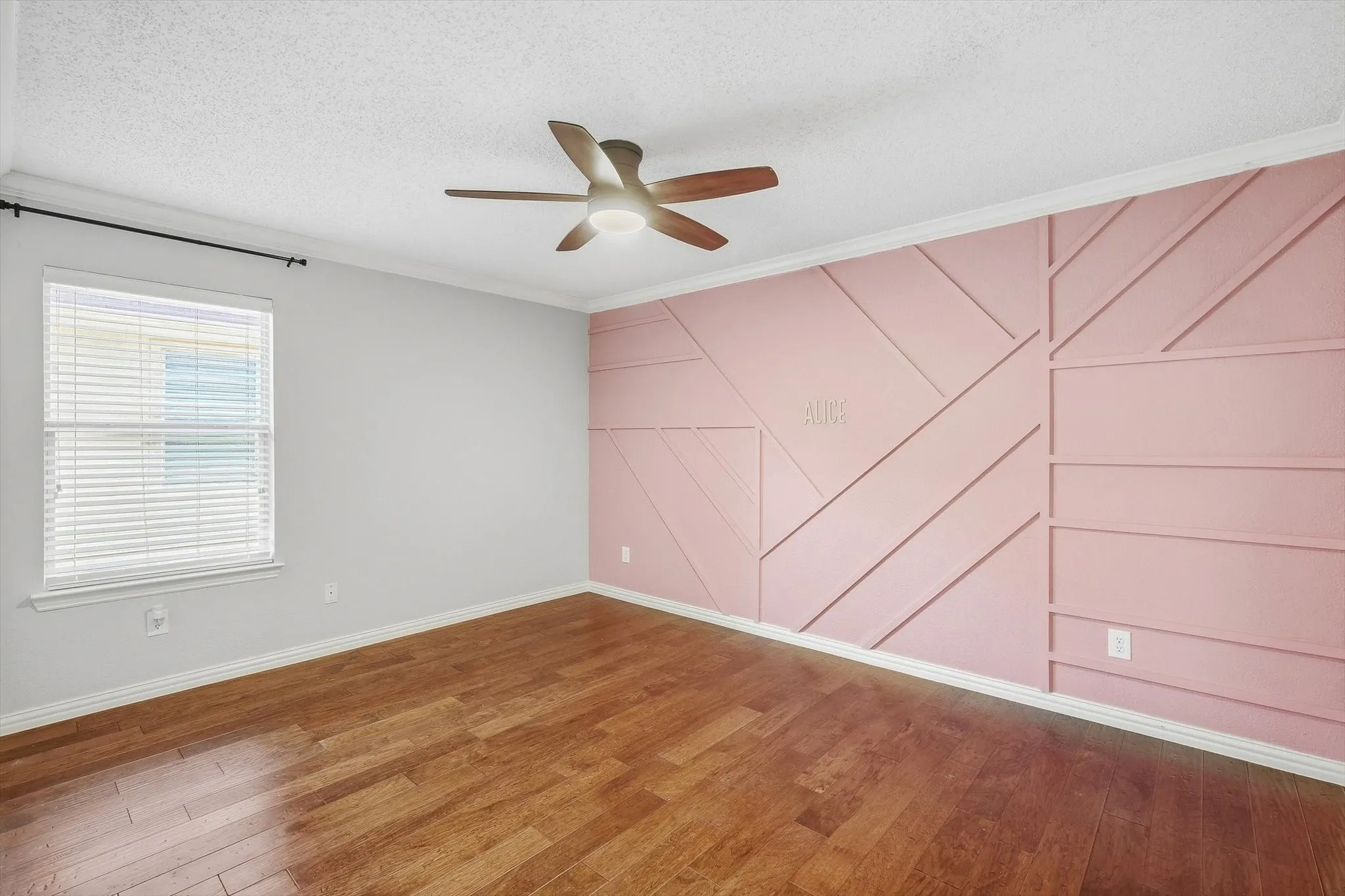 Wood-finish flooring, accent wall with geometric paneling, ceiling fan with integrated lighting, window with horizontal blinds, and white trim molding