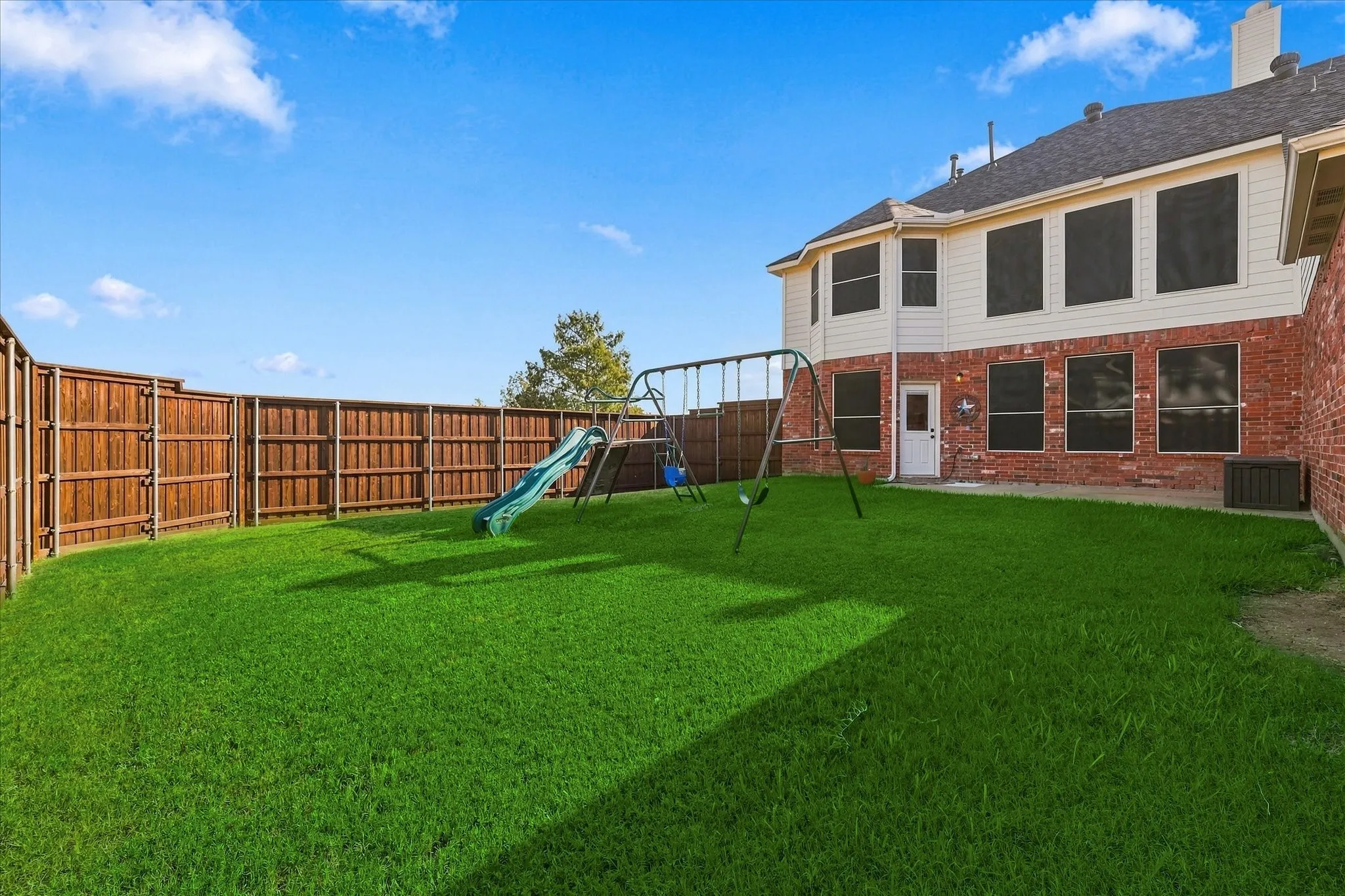 Expansive grassy backyard featuring a full privacy fence, brick exterior, and a white access door