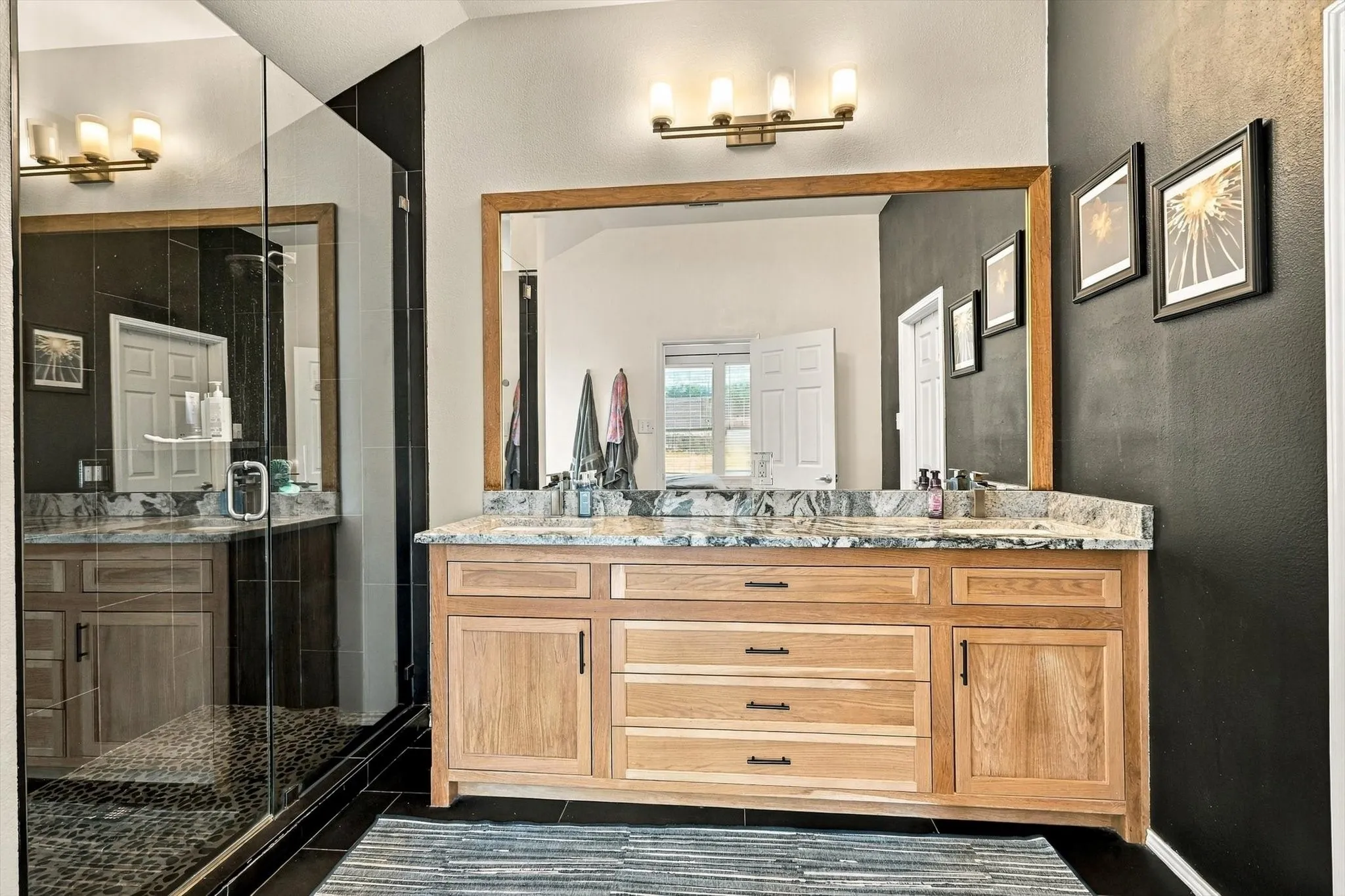 Dual basin vanity with natural wood-finish cabinetry, stone countertop, framed mirror, and glass-enclosed shower