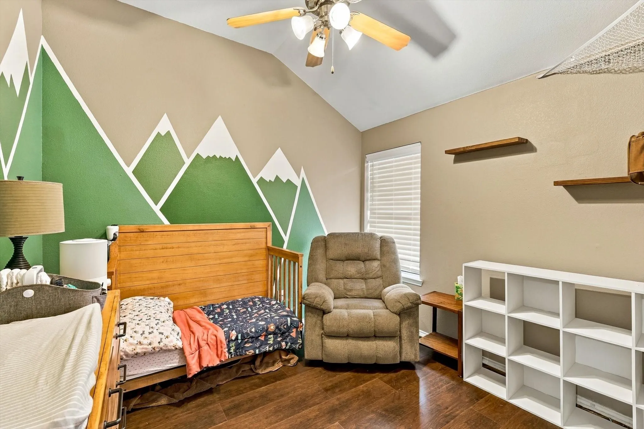 Vaulted ceiling with a ceiling fan, wood-finish flooring, accent wall with geometric mountain design, window with blinds, and built-in wall shelving