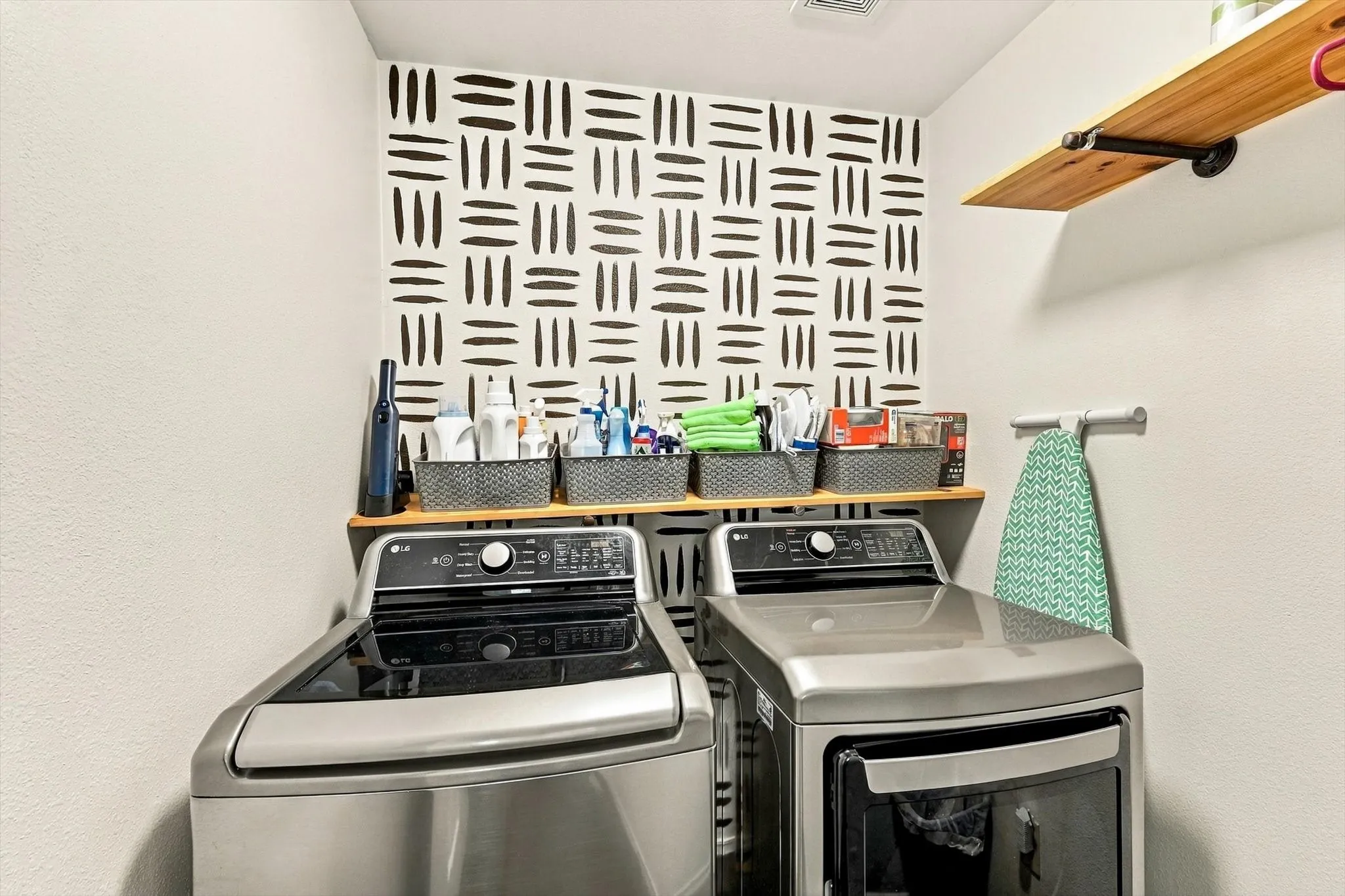 Laundry area with a patterned accent wall, two top-load appliances, and a wood-finish floating shelf