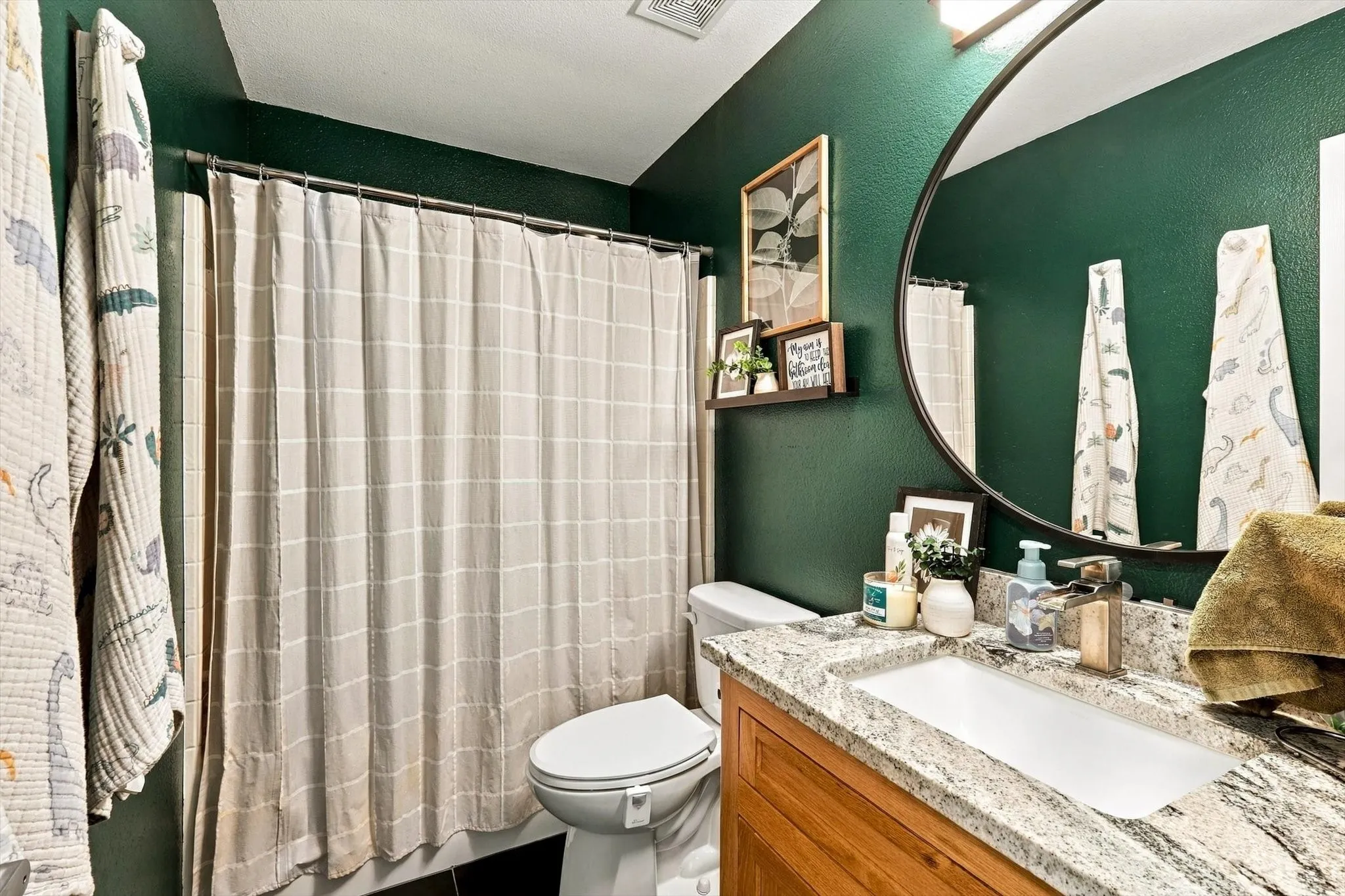Granite-style countertop with integrated sink, wood-finish vanity, round framed mirror, and a shower with a patterned curtain
