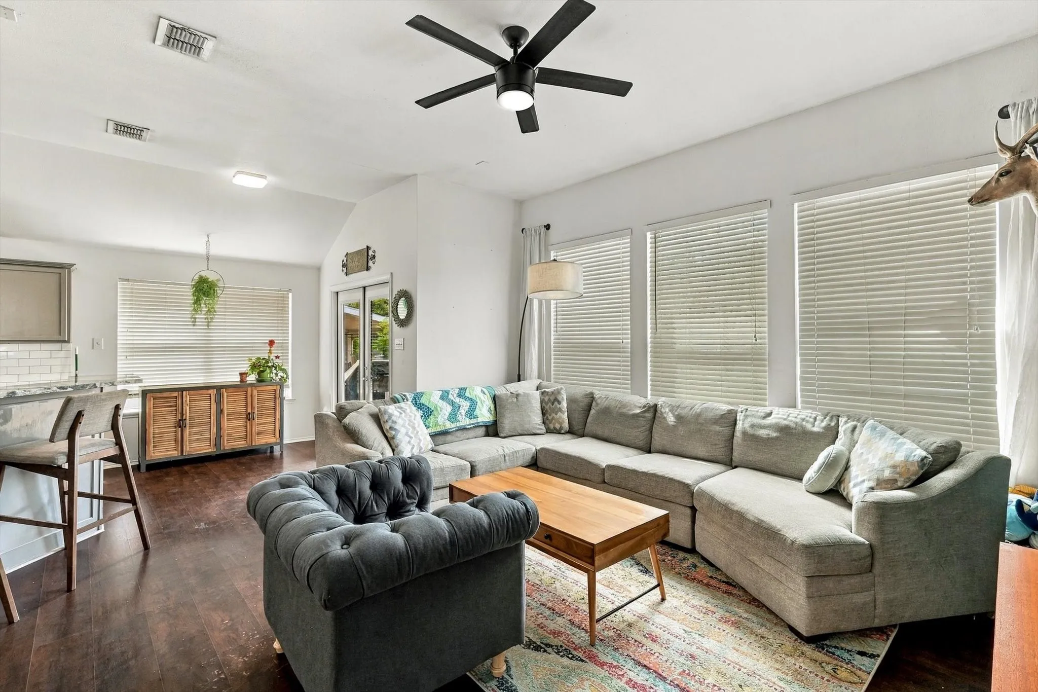 Vaulted ceilings, dark wood-finish flooring, multiple windows with blinds, a ceiling fan with integrated lighting, and an open-concept layout