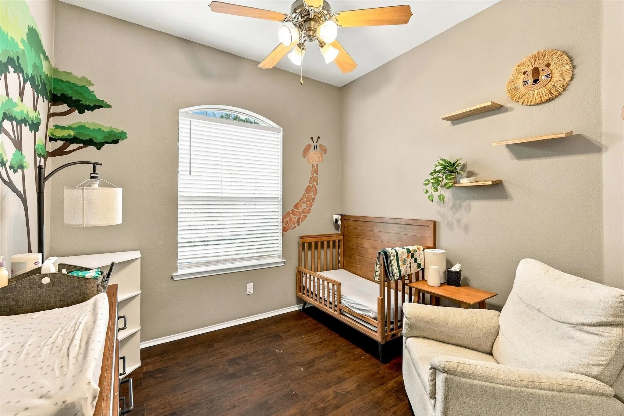 Wood-finish flooring, ceiling fan with integrated lighting, arched window with horizontal blinds, and a built-in wall shelf system