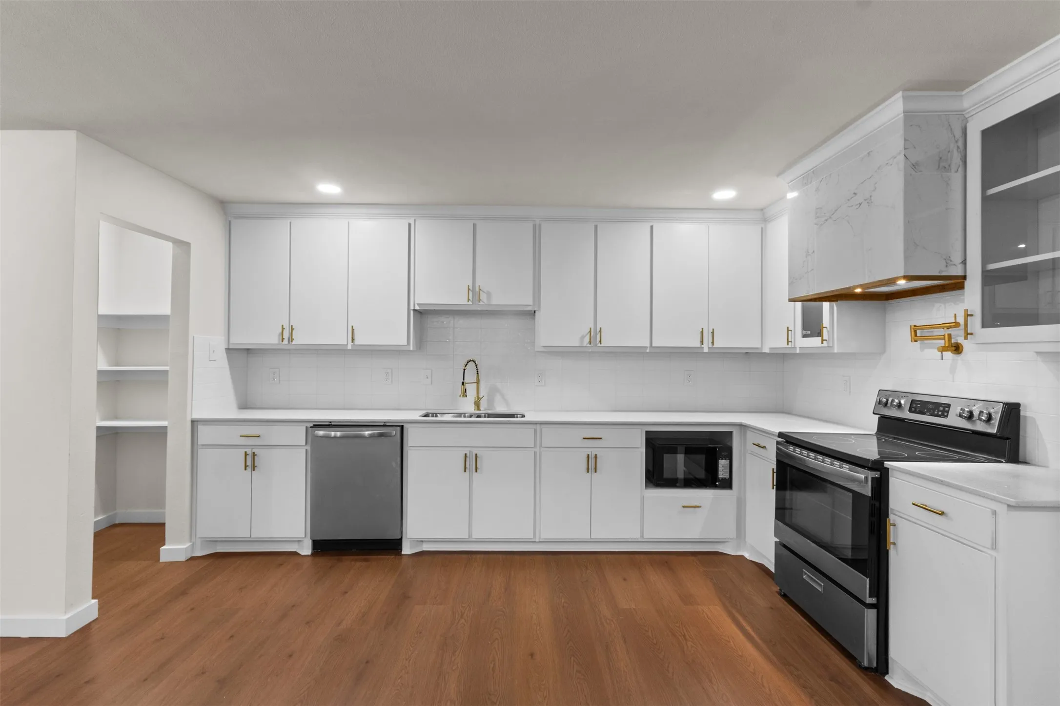 White shaker-style cabinetry with gold-tone hardware, wood-finish flooring, undermount sink with a gold-tone faucet, integrated stainless steel dishwasher, and a built-in microwave