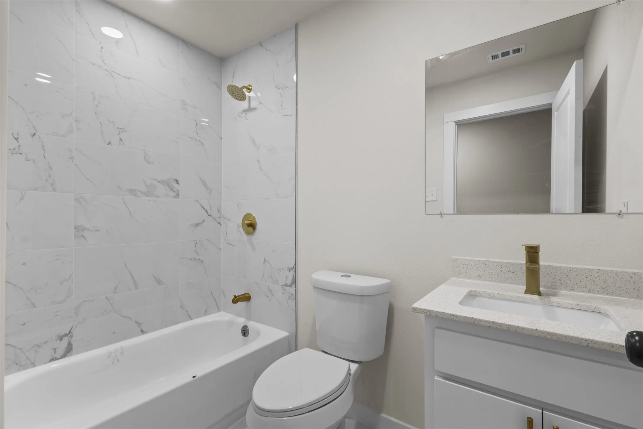 Bathtub with white marble-look tile surround, gold-finish fixtures, and a white vanity with a light-colored speckled countertop